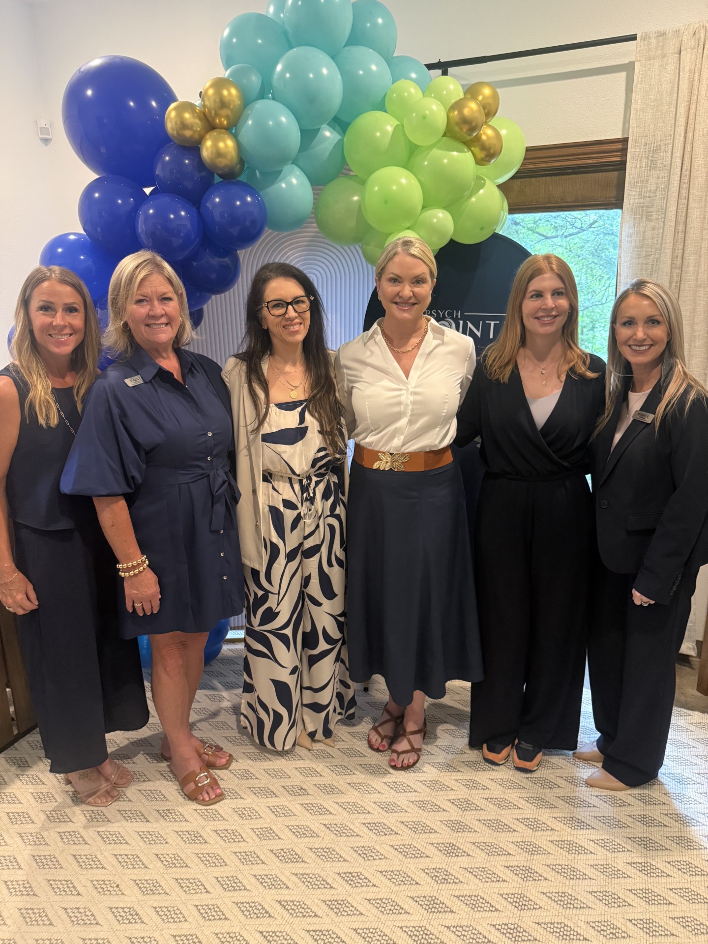 Six women standing indoors in front of colorful balloon arrangement, smiling, dressed in formal and semi-formal attire.