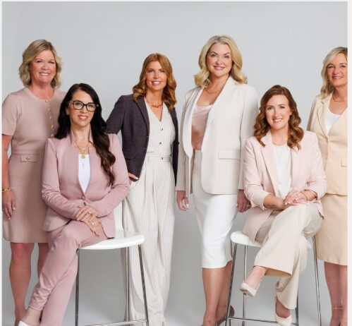 Six women in professional attire pose together against a light gray background.