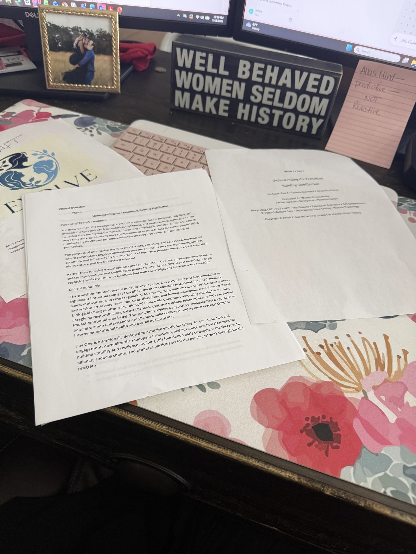 Desk with open papers, a floral tablecloth, a framed photo, and a sign reading 'Well behaved women seldom make history.'