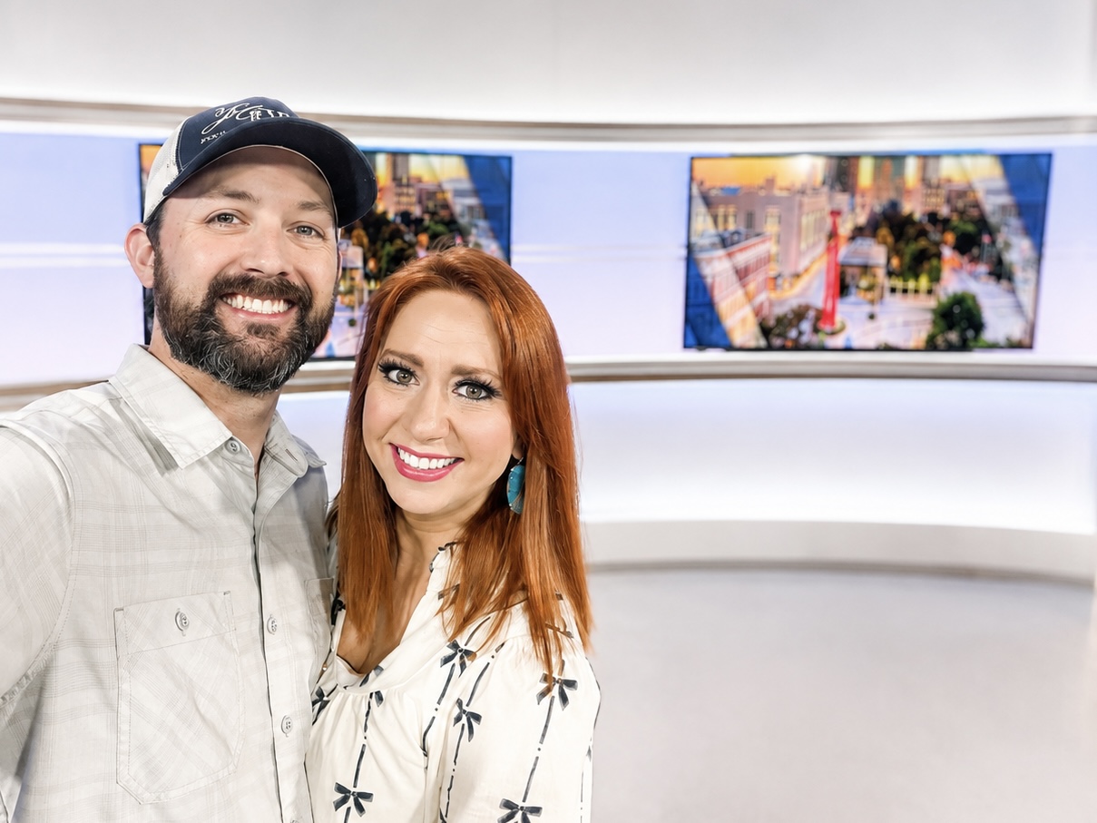 Two smiling people, a man with a beard and a woman with red hair, stand in front of screens displaying cityscapes.