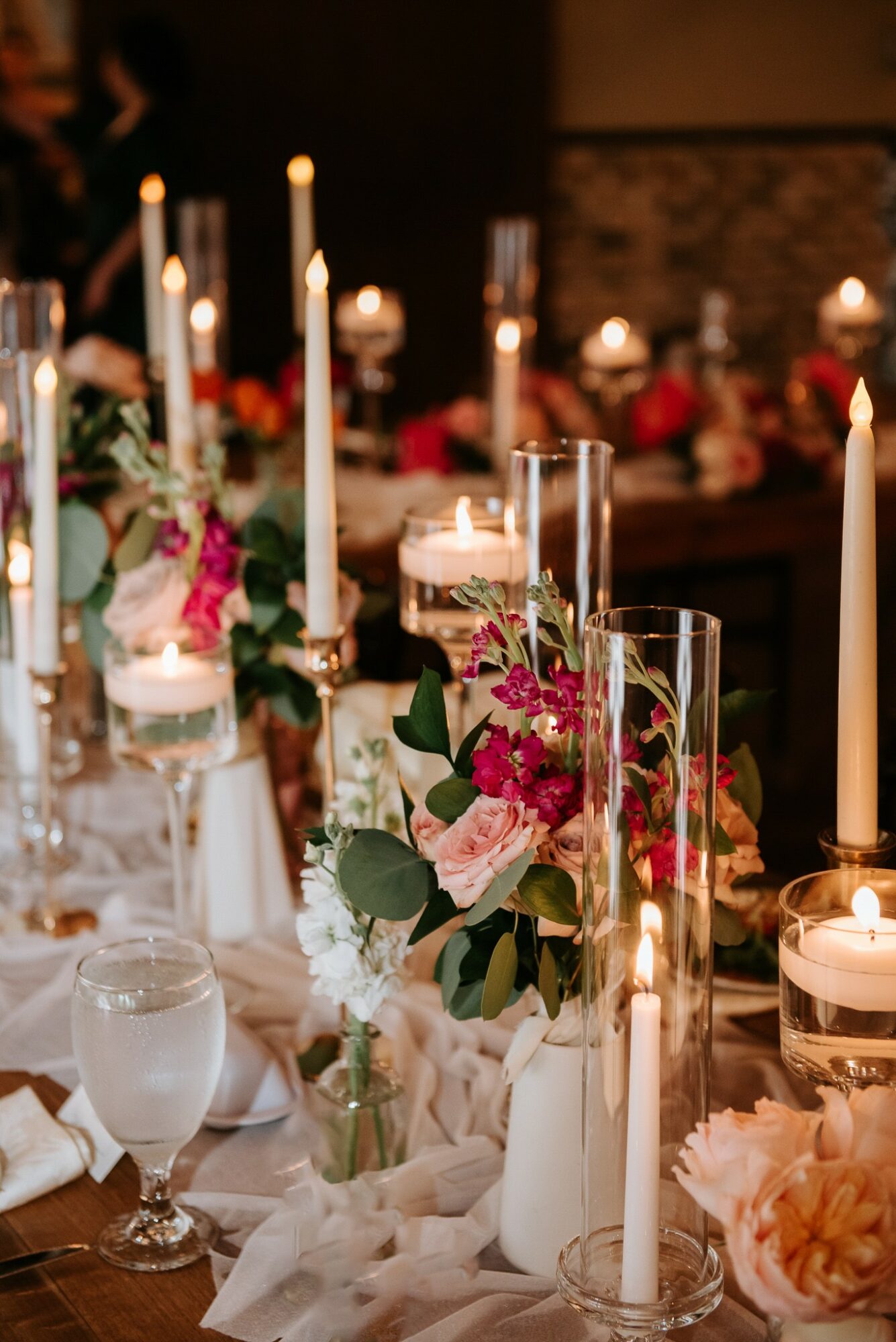 Elegant table setting with tall candles, floral arrangements, and glassware in a dimly lit room.