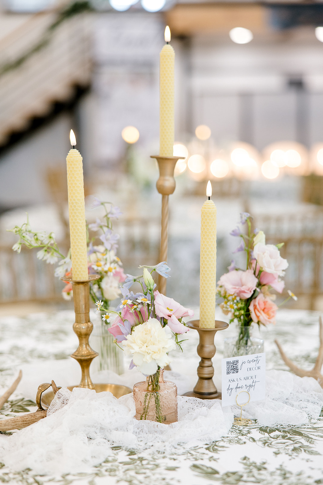 Three tall candles in ornate holders with floral arrangements on a table, blurred background, soft lighting.
