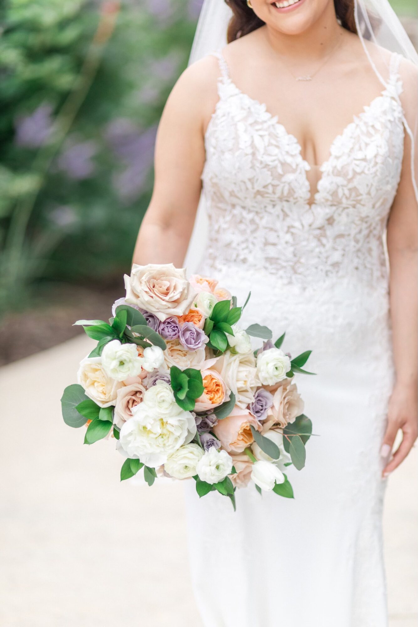 Bride in a lace wedding dress holding a bouquet of roses and greenery, outdoors with blurred greenery background.