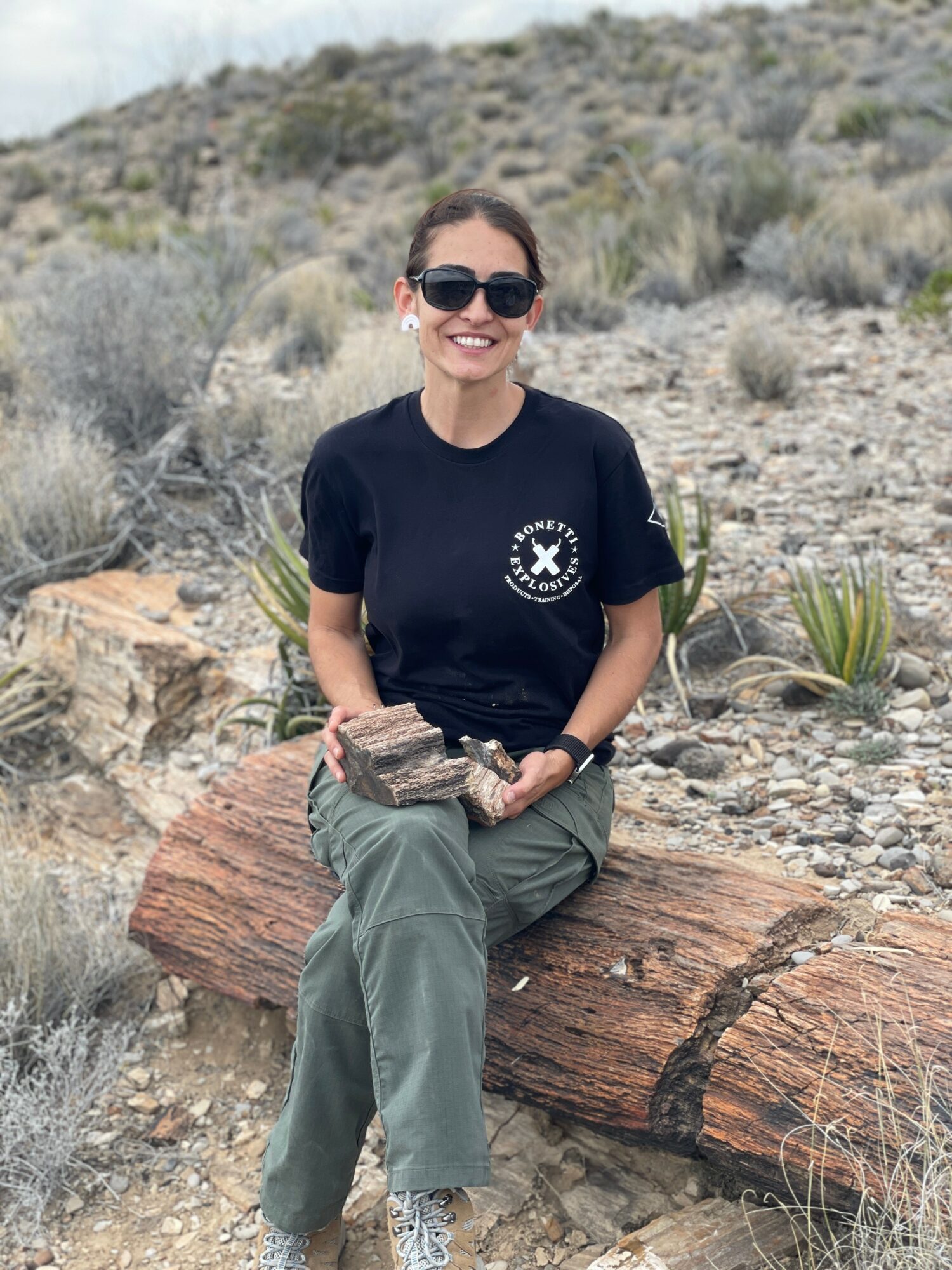 Woman sitting on a log in a desert landscape, smiling, wearing sunglasses, a black t-shirt, and green pants.
