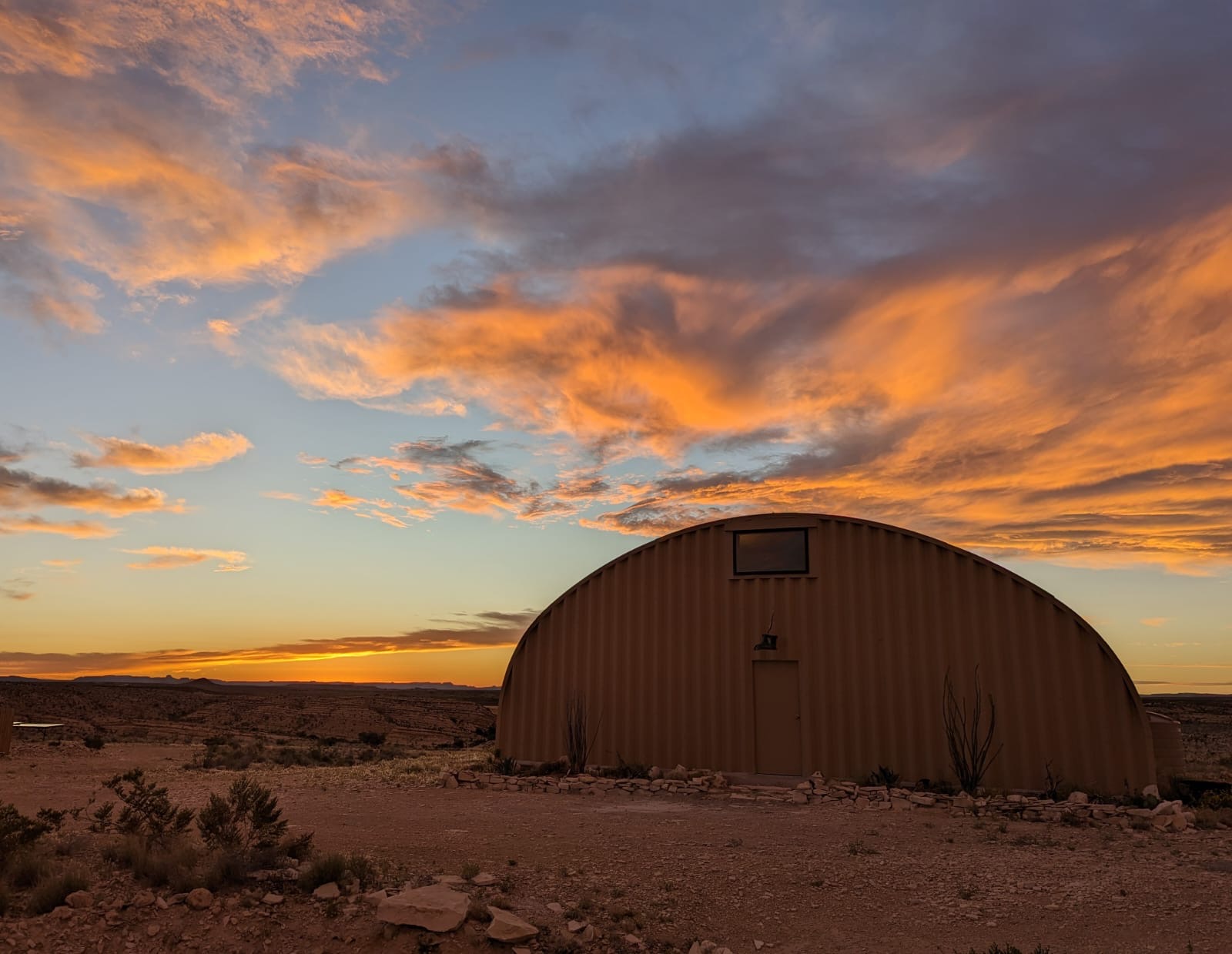 A large, curved metal building in a desert landscape during sunset with colorful clouds in the sky.