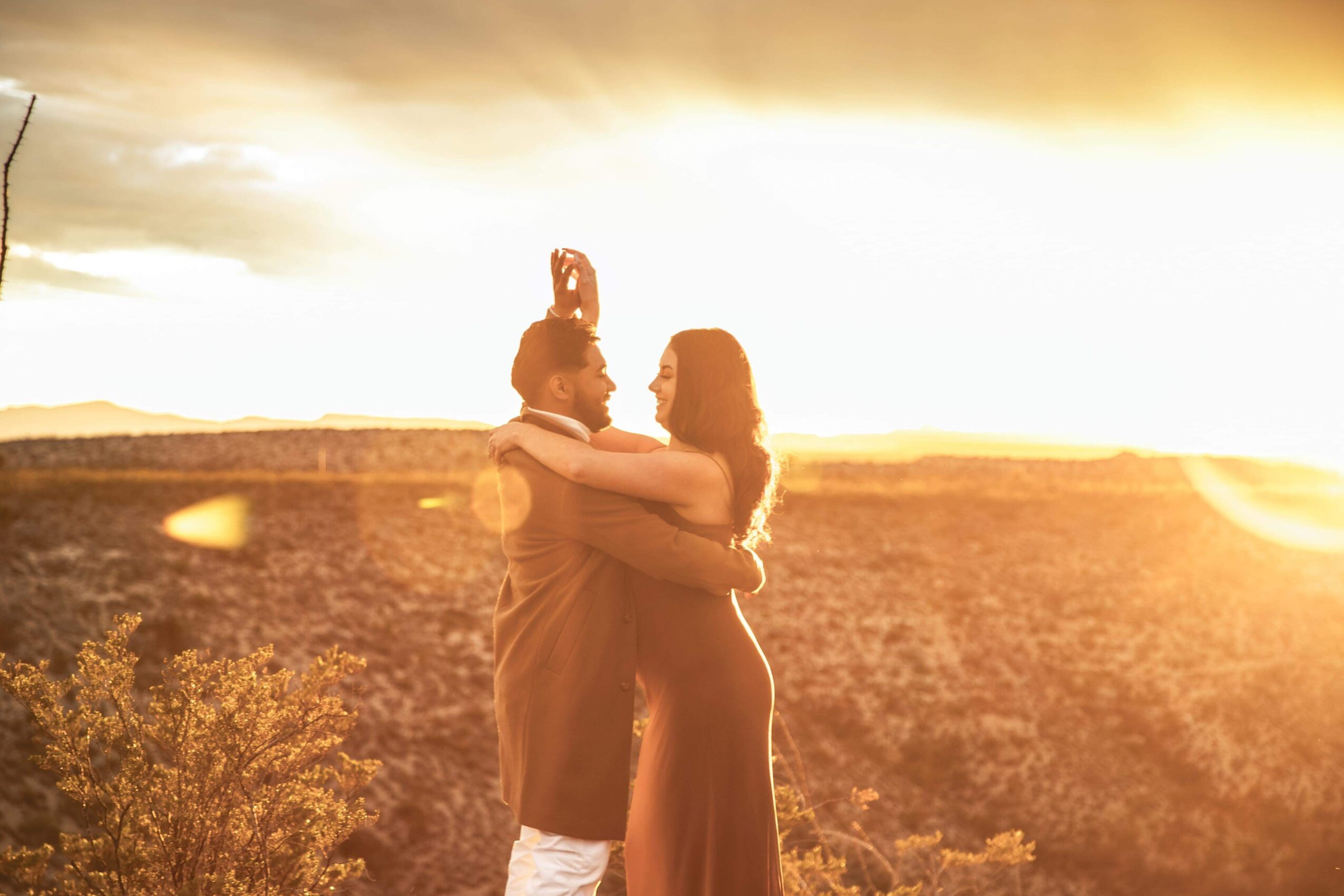Two people embrace outdoors during sunset, with a landscape and sky in the background, warm lighting, and lens flare effects.