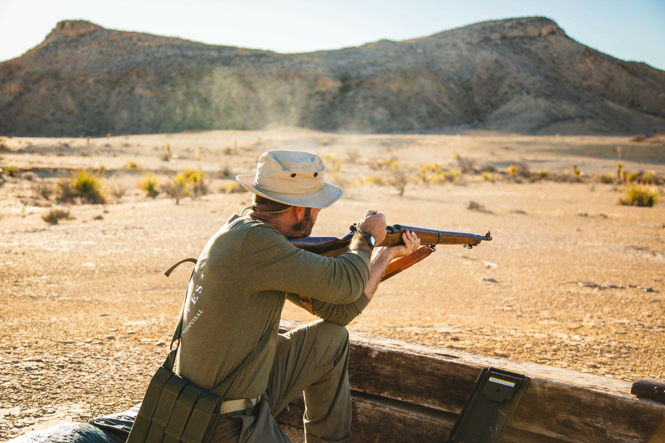 Person in outdoor desert landscape aiming rifle, wearing hat and green clothing, with mountains in background.