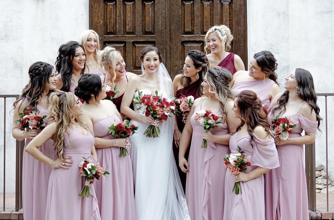 Group of women in pink dresses and a bride in white dress holding bouquets, standing outside a building with wooden door.