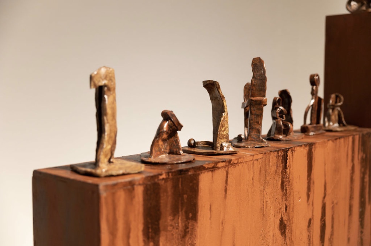 Small metallic sculptures resembling abstract figures are displayed on a wooden surface, with a plain background.