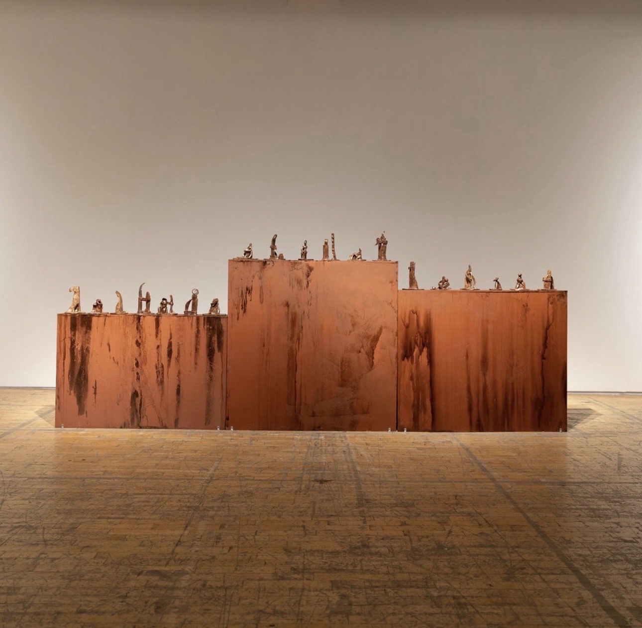 Three rust-colored rectangular blocks with small sculptures on top, set against a plain wall and wooden floor.