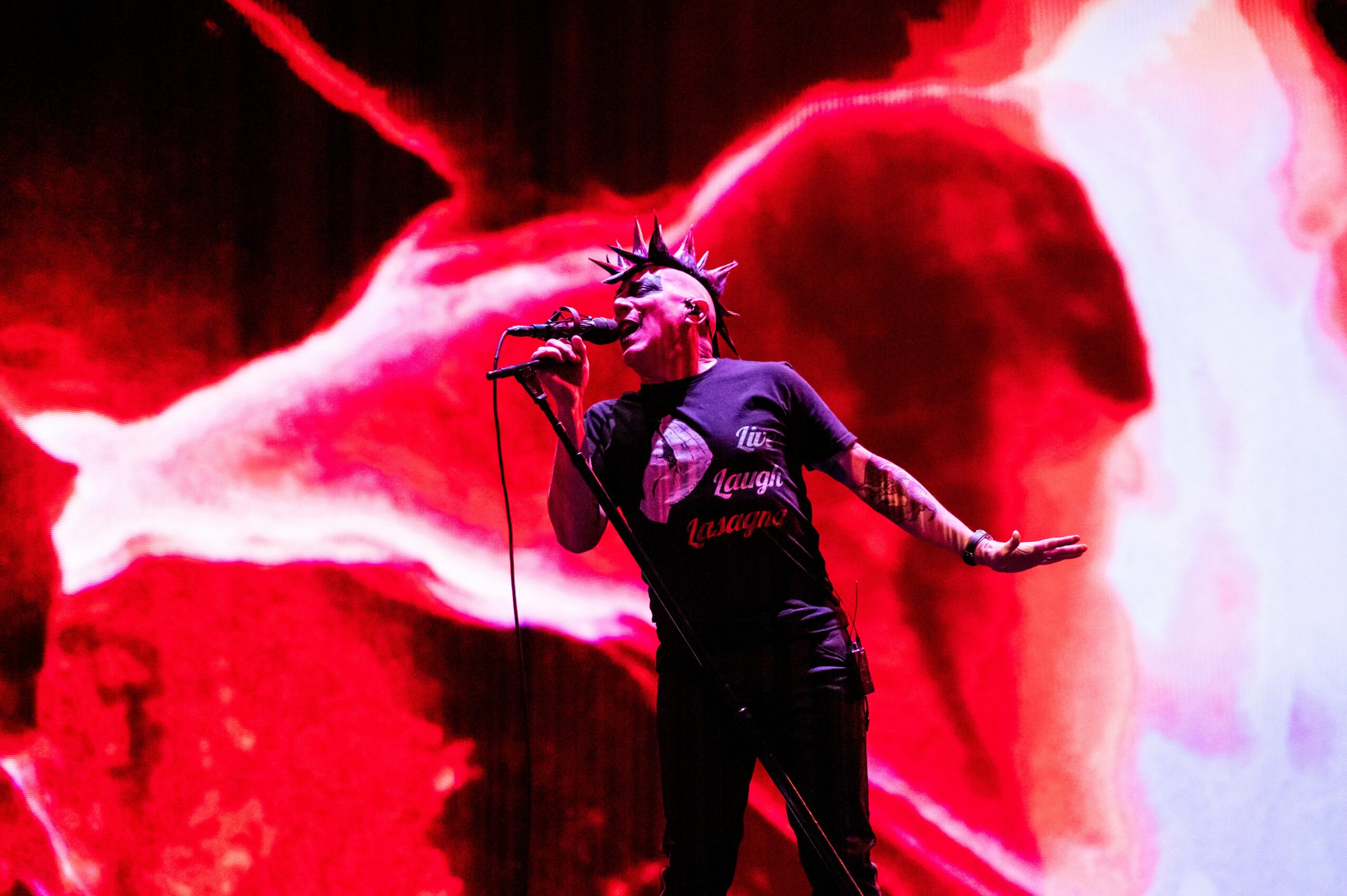 Performer with spiked hair singing into a microphone on stage with a red and white abstract background.