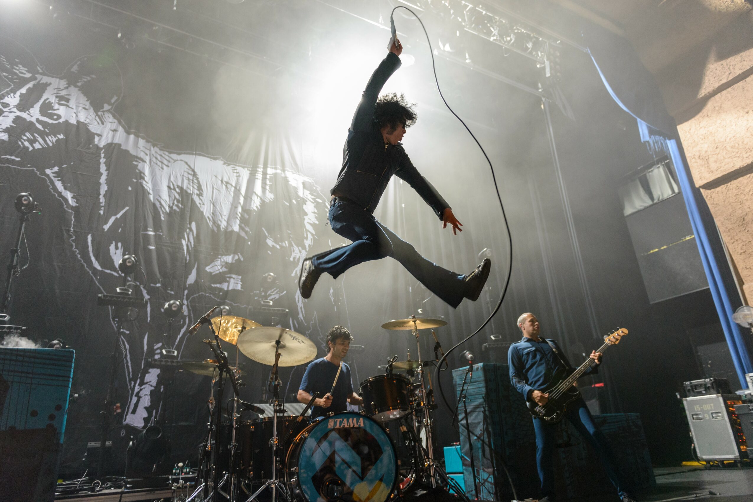 A band performs on stage with a singer jumping in the air holding a microphone cord, drums, and guitarists visible.