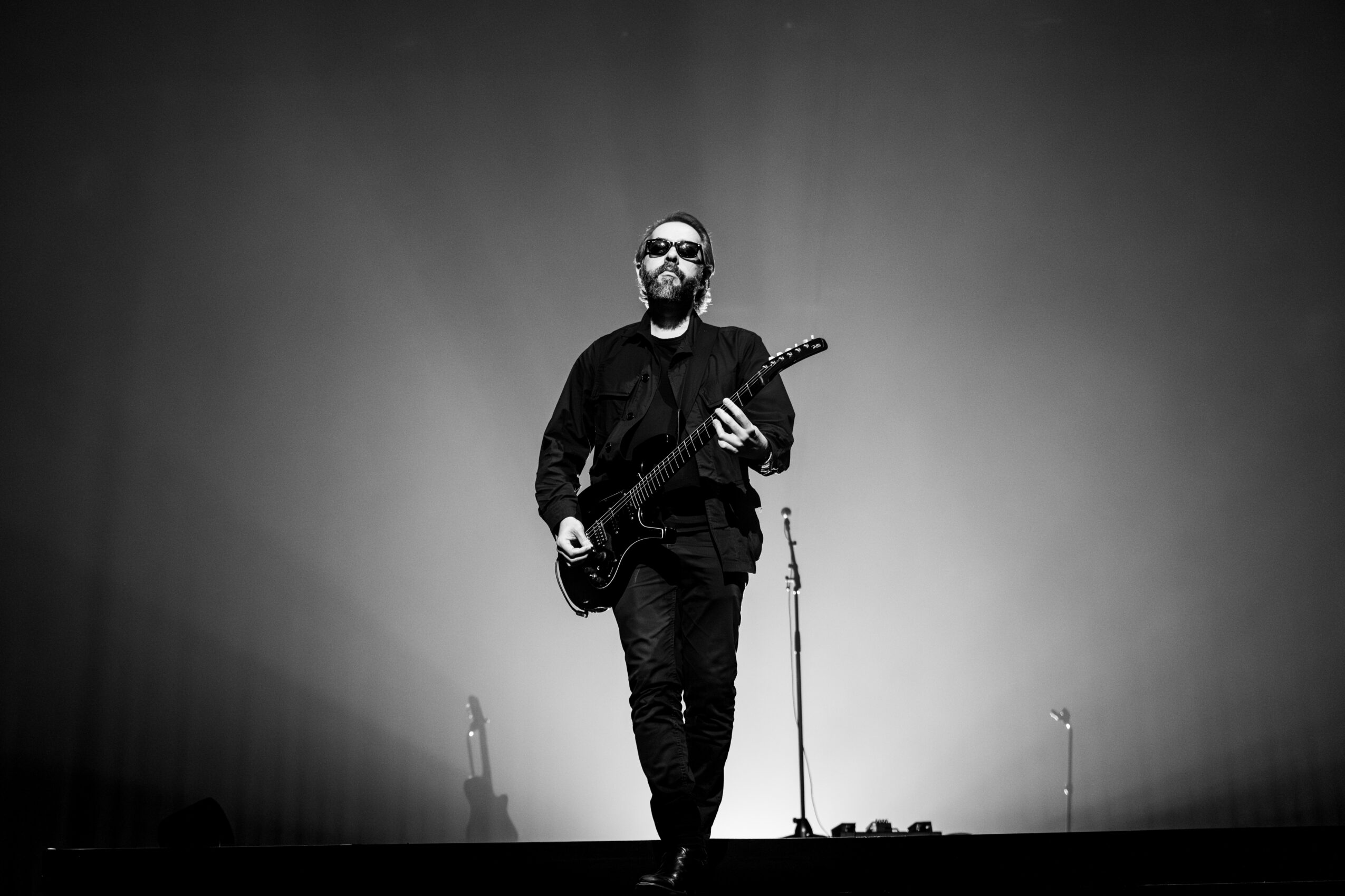 Man with sunglasses holding a guitar on stage, microphone stand nearby, in black and white, with a plain background.