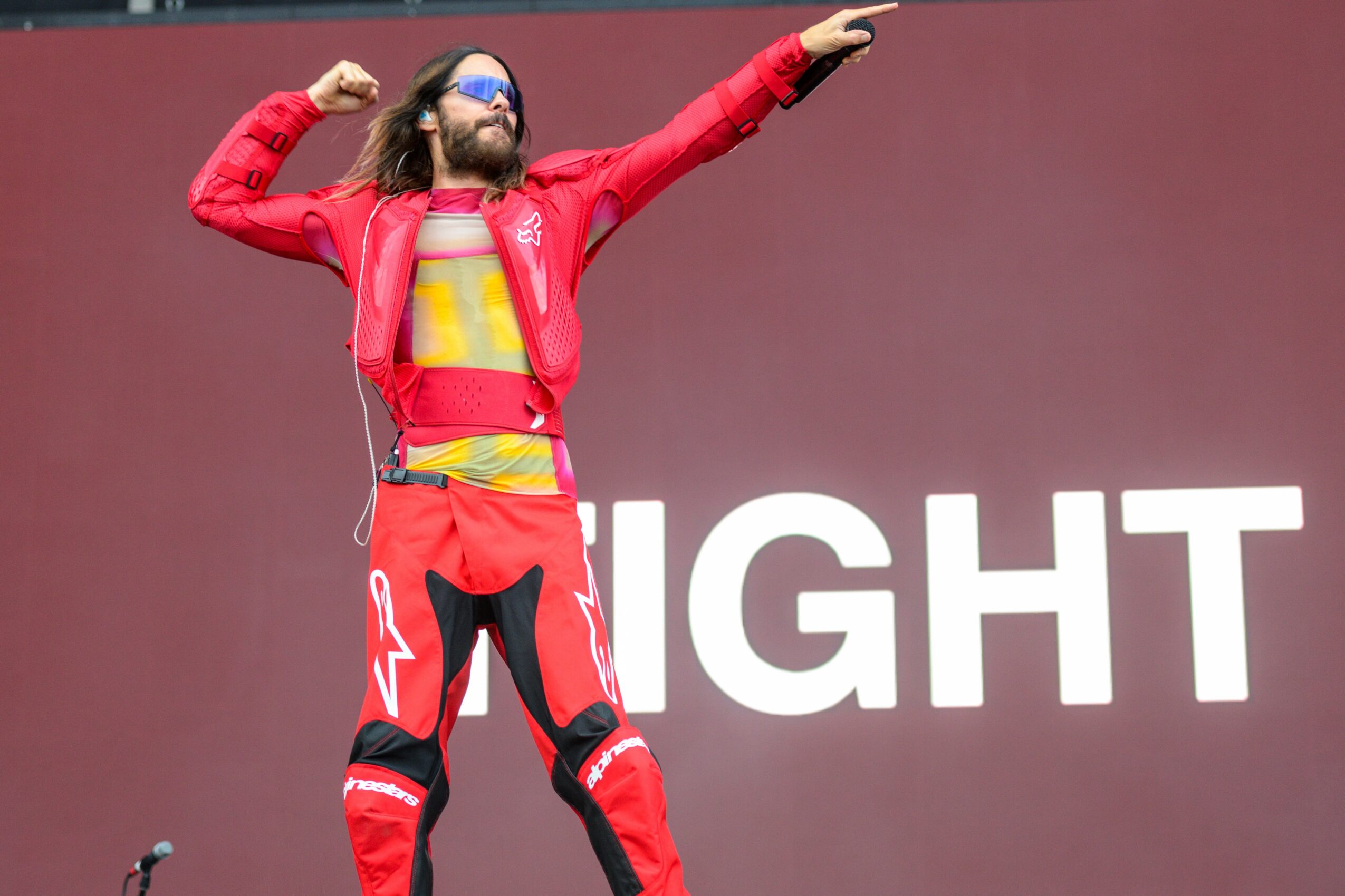 Person with long hair and beard wearing red outfit and sunglasses, raising one arm, standing in front of a red background with white text.