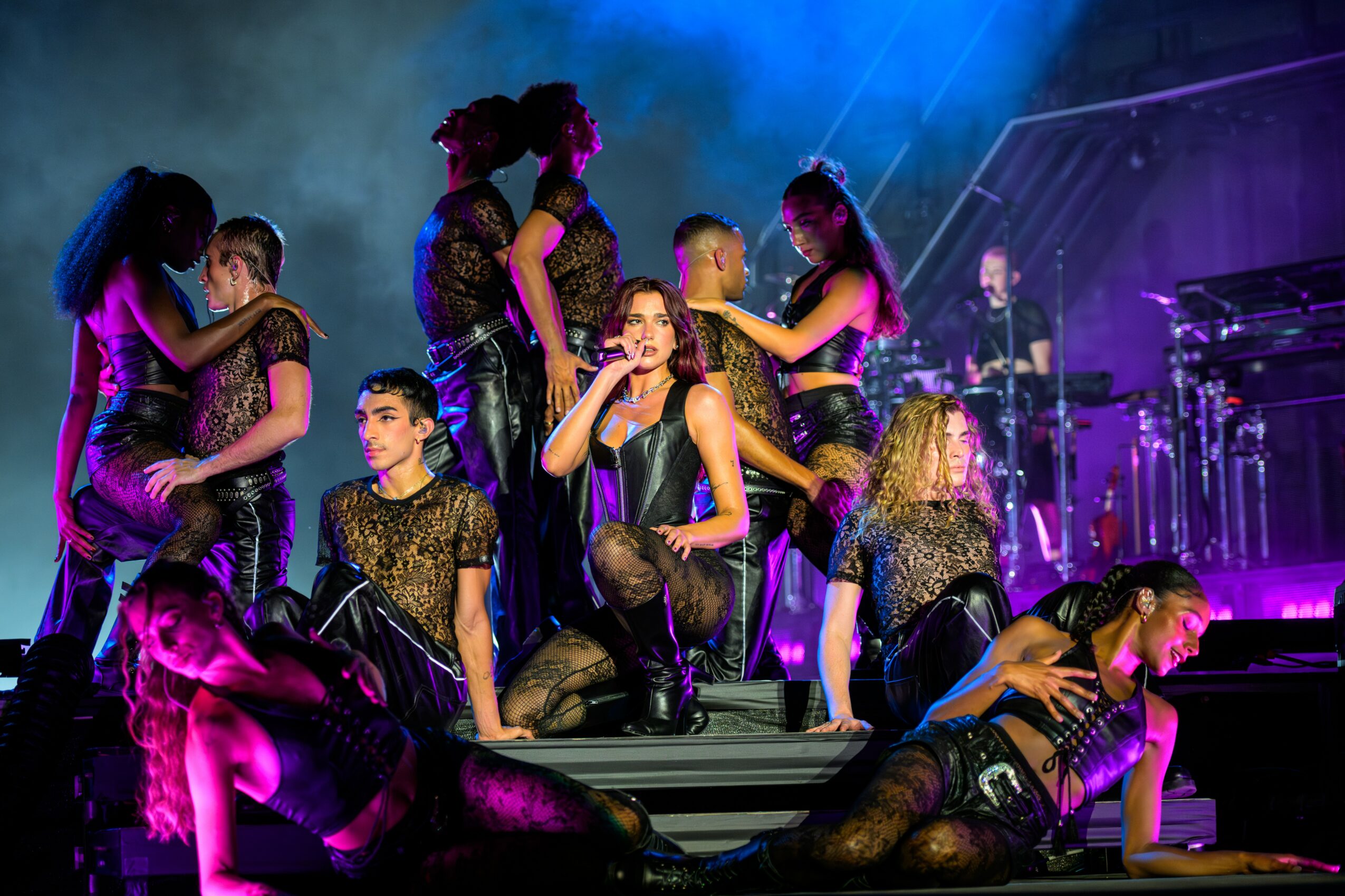 Group of performers on stage, some sitting and others standing, with a band playing in the background, colorful stage lighting.