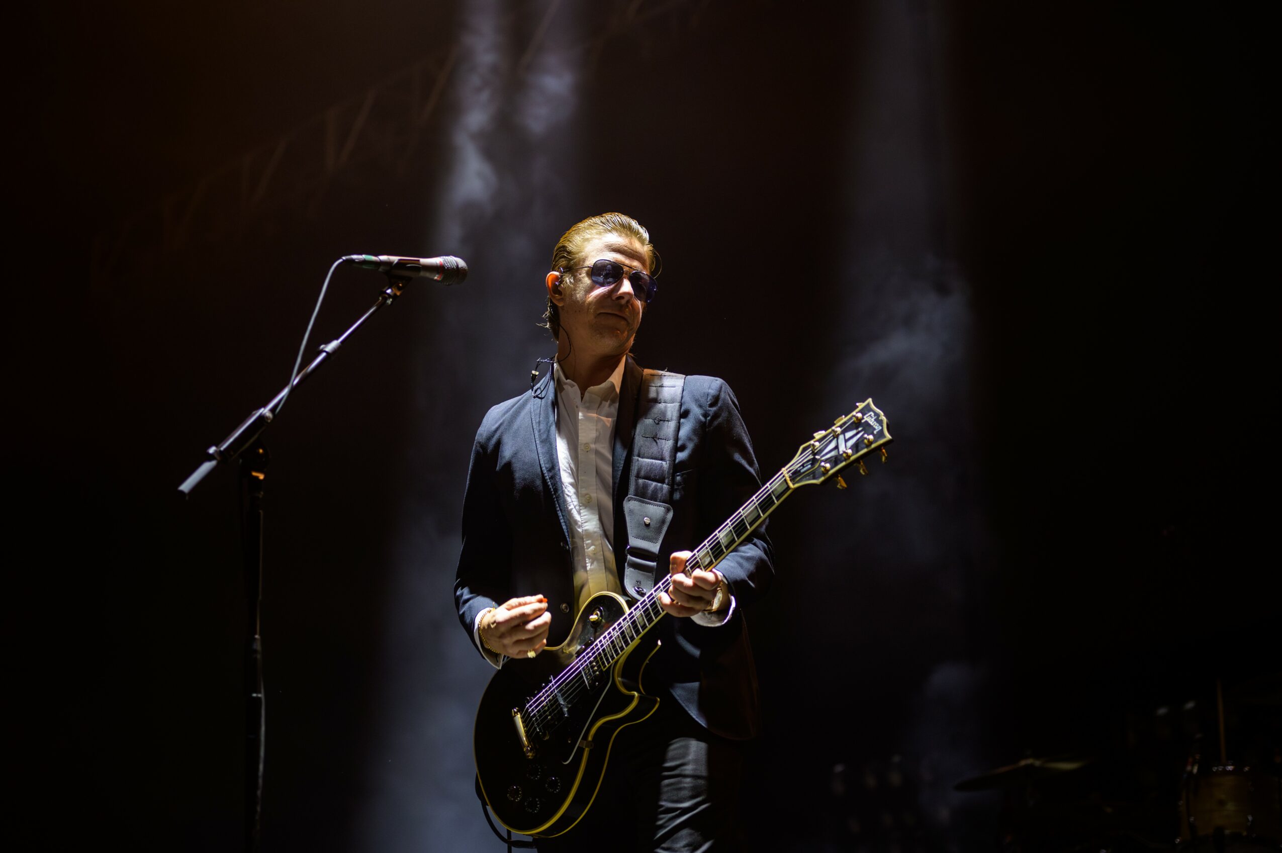 Person playing electric guitar on stage with dark background and spotlight, wearing sunglasses and a dark suit.