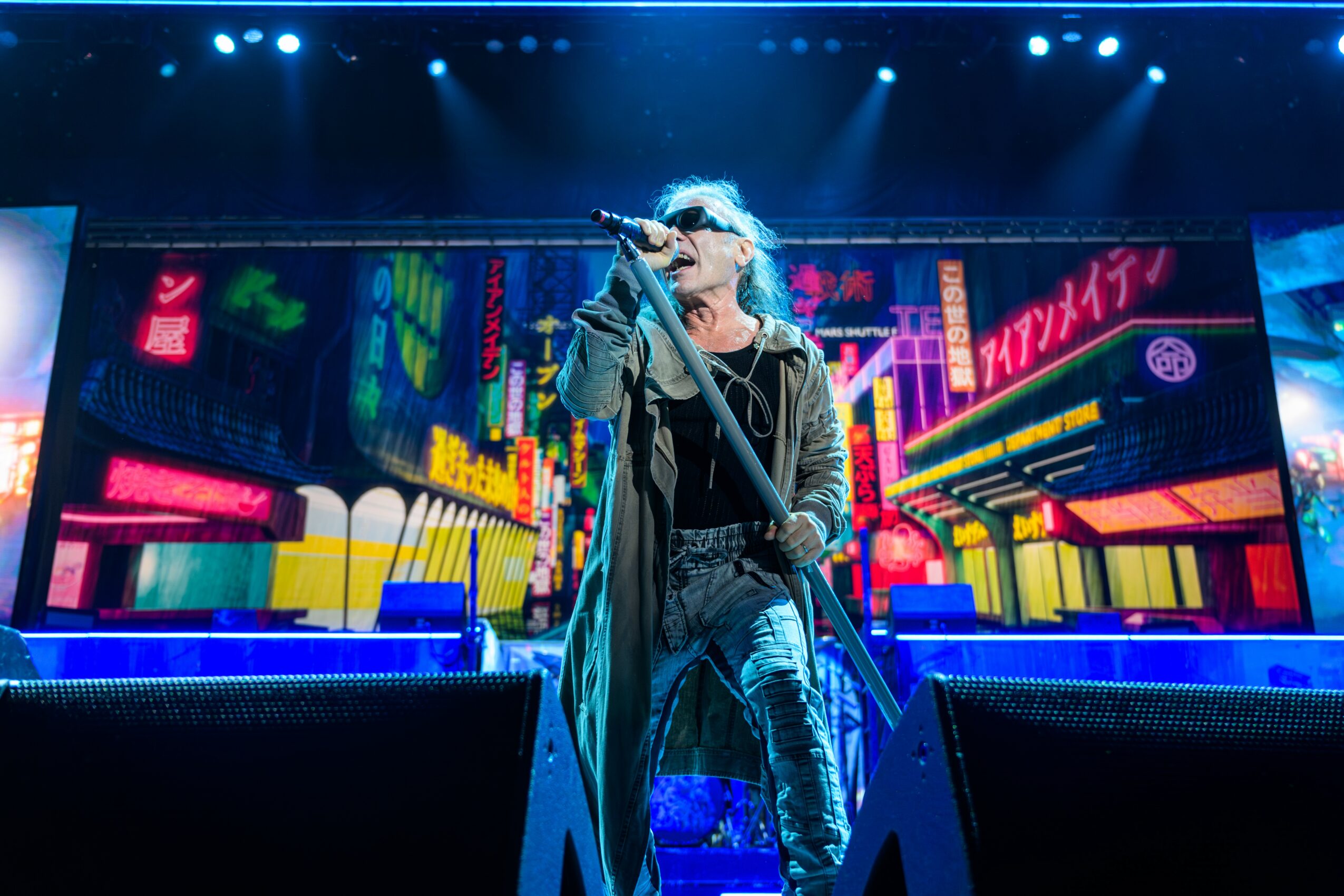 Performer singing on stage with colorful cityscape background, holding a microphone, wearing sunglasses and casual clothing.