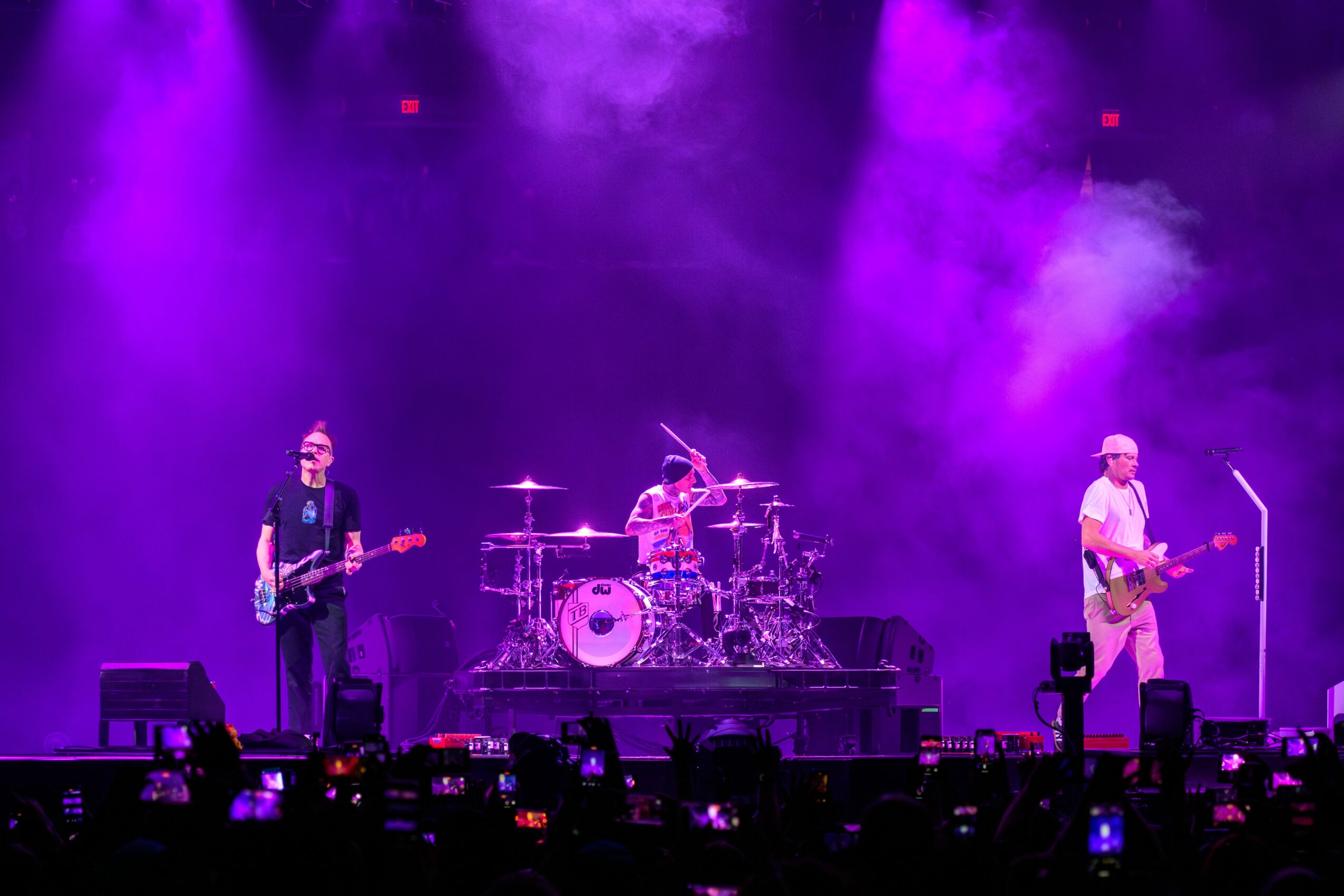 Three musicians perform on stage with purple lighting and smoke, playing guitars and drums, audience capturing the moment.
