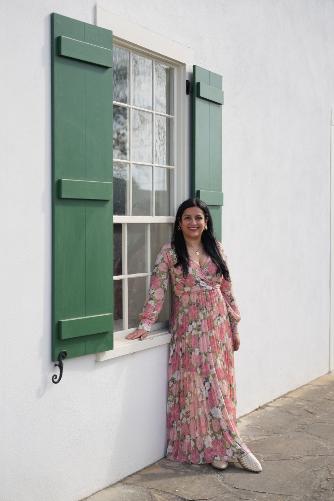 Woman in floral dress standing outside near a window with green shutters, white wall, and stone ground.