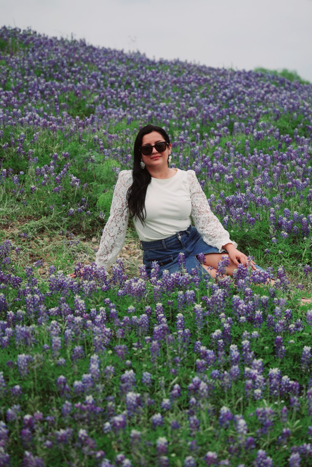Woman sitting in a field of purple flowers, wearing sunglasses, white top, and denim shorts, smiling at the camera.