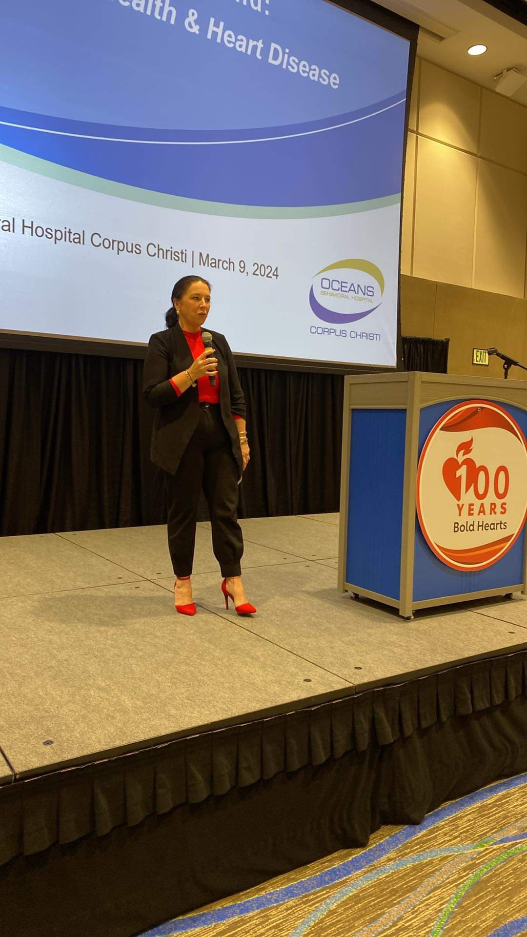 Woman speaking on stage with a presentation slide behind her, standing near a podium with a heart logo, in a conference room.