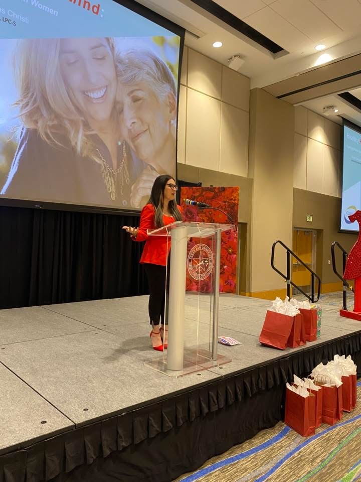 Woman speaking at a podium on stage with large screen showing a photo of two smiling women behind her.