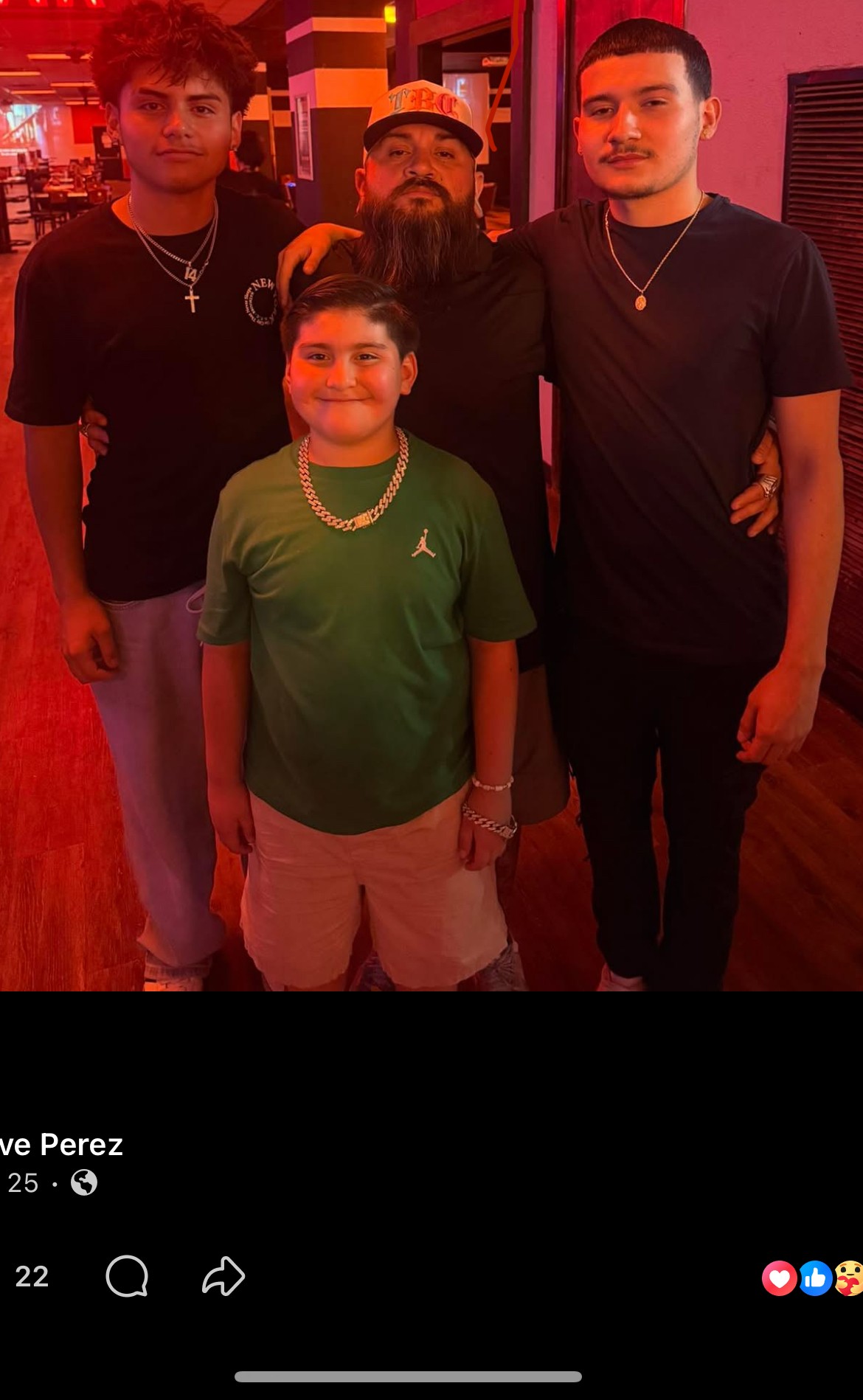 Four people standing together indoors, three young men and one boy, with colorful lighting in the background.
