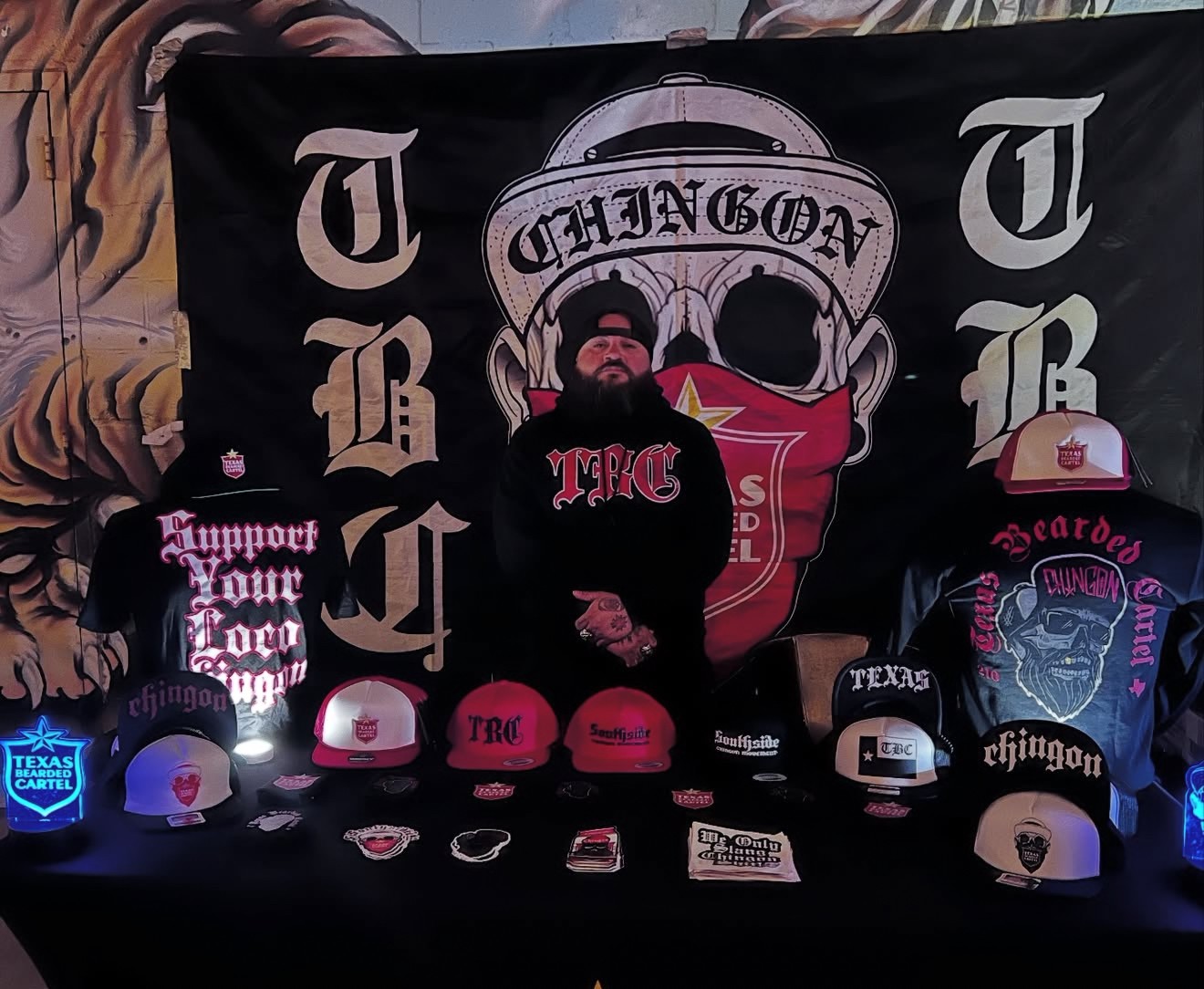 Person wearing a black hoodie with Texas flag, surrounded by hats and patches, in front of a large Texas-themed banner.