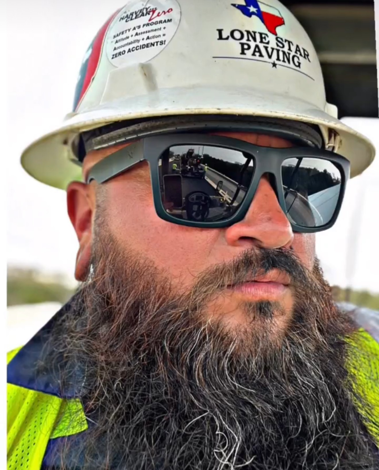Construction worker with sunglasses, hard hat, and beard, wearing a high-visibility vest, outdoors.
