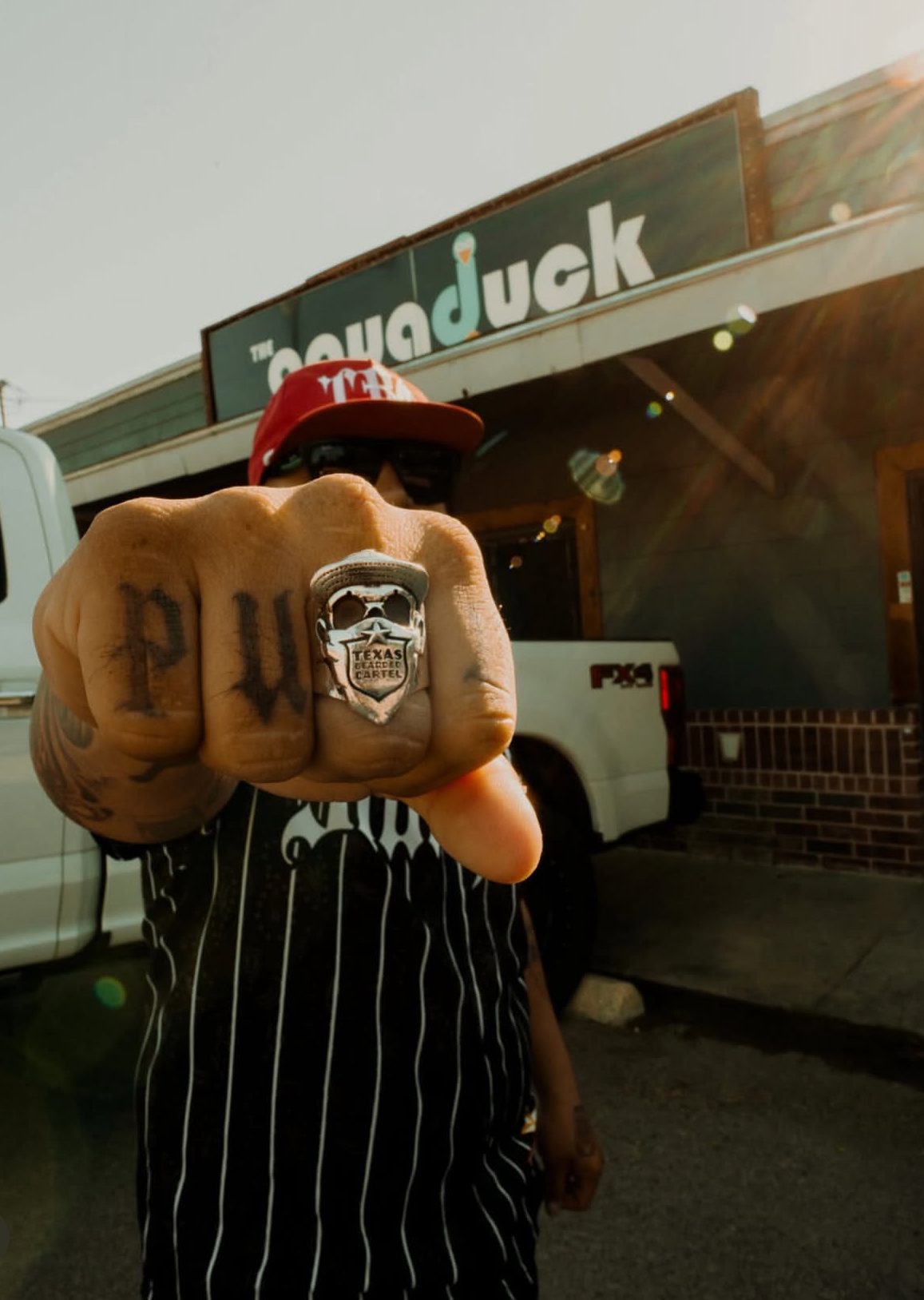 Person wearing a striped shirt and red cap shows a fist with a skull tattoo and a skull ring, standing outside a building.