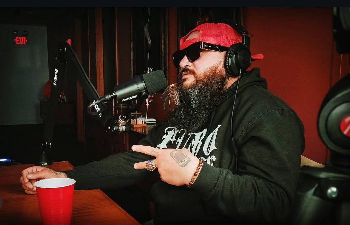 Man with beard, sunglasses, red cap, and headphones speaking into a microphone in a recording studio.