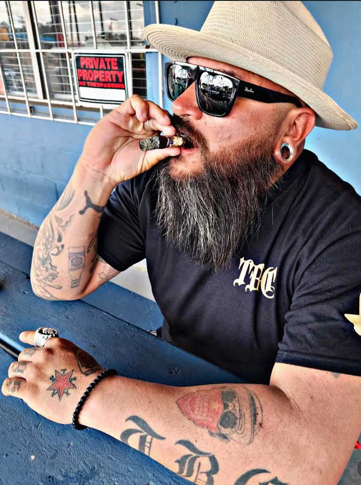 Man with a beard, sunglasses, hat, and tattoos, eating a snack outdoors, sitting at a blue table.