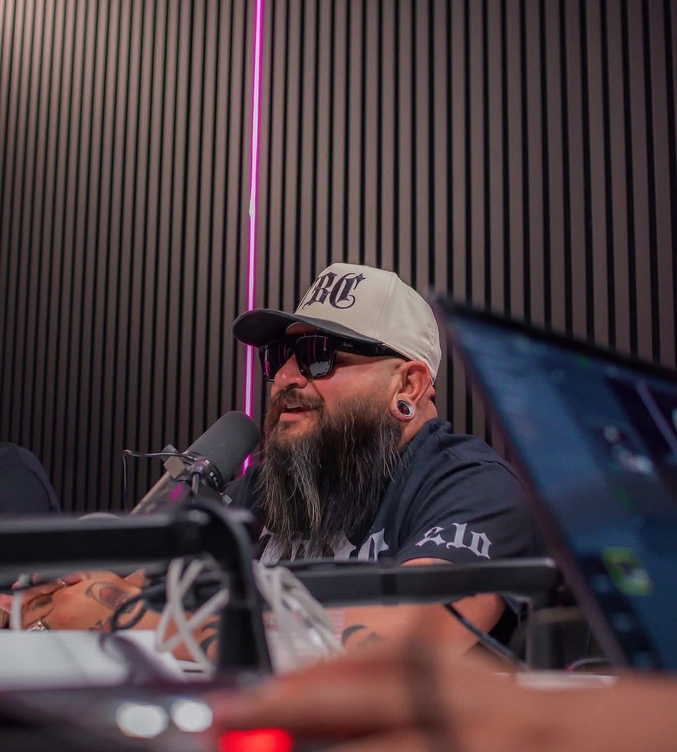 Man with a beard, sunglasses, and a cap speaking into a microphone in a recording studio with black soundproof panels.