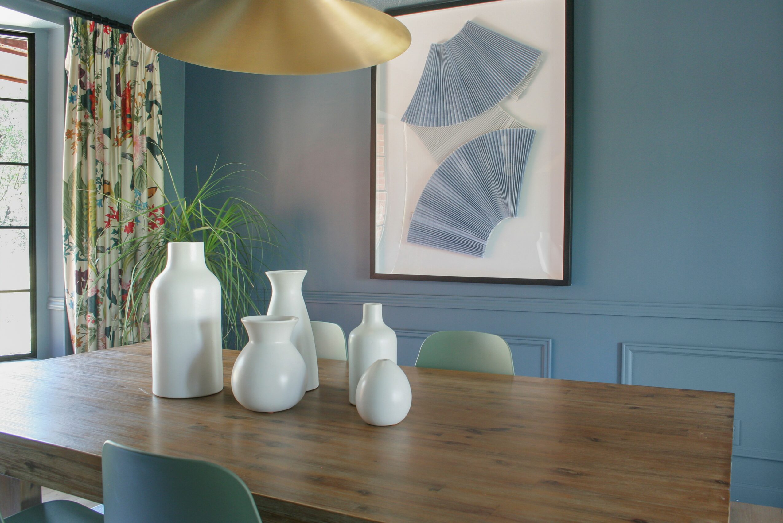 Wooden dining table with white vases, blue wall with framed artwork, and a gold pendant light above.
