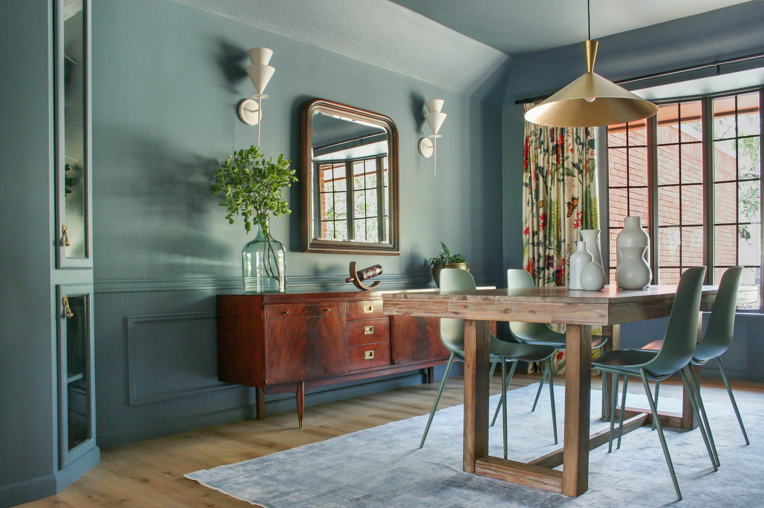 Dining room with a wooden table, six chairs, a sideboard, mirror, and large window with floral curtains.