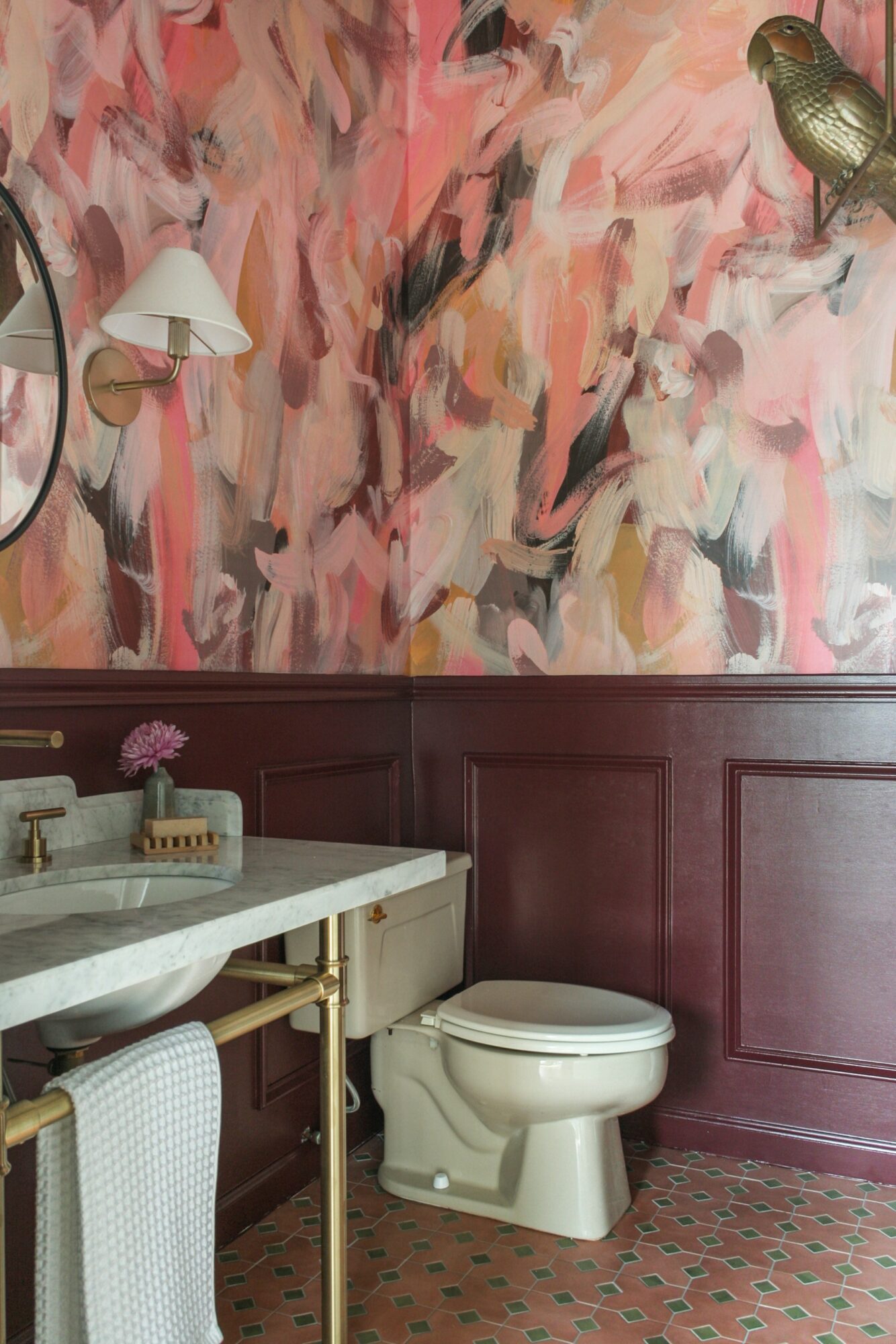 Bathroom with pink and beige abstract wall art, white sink, toilet, and patterned floor, with wall-mounted lamp and decorative bird sculpture.