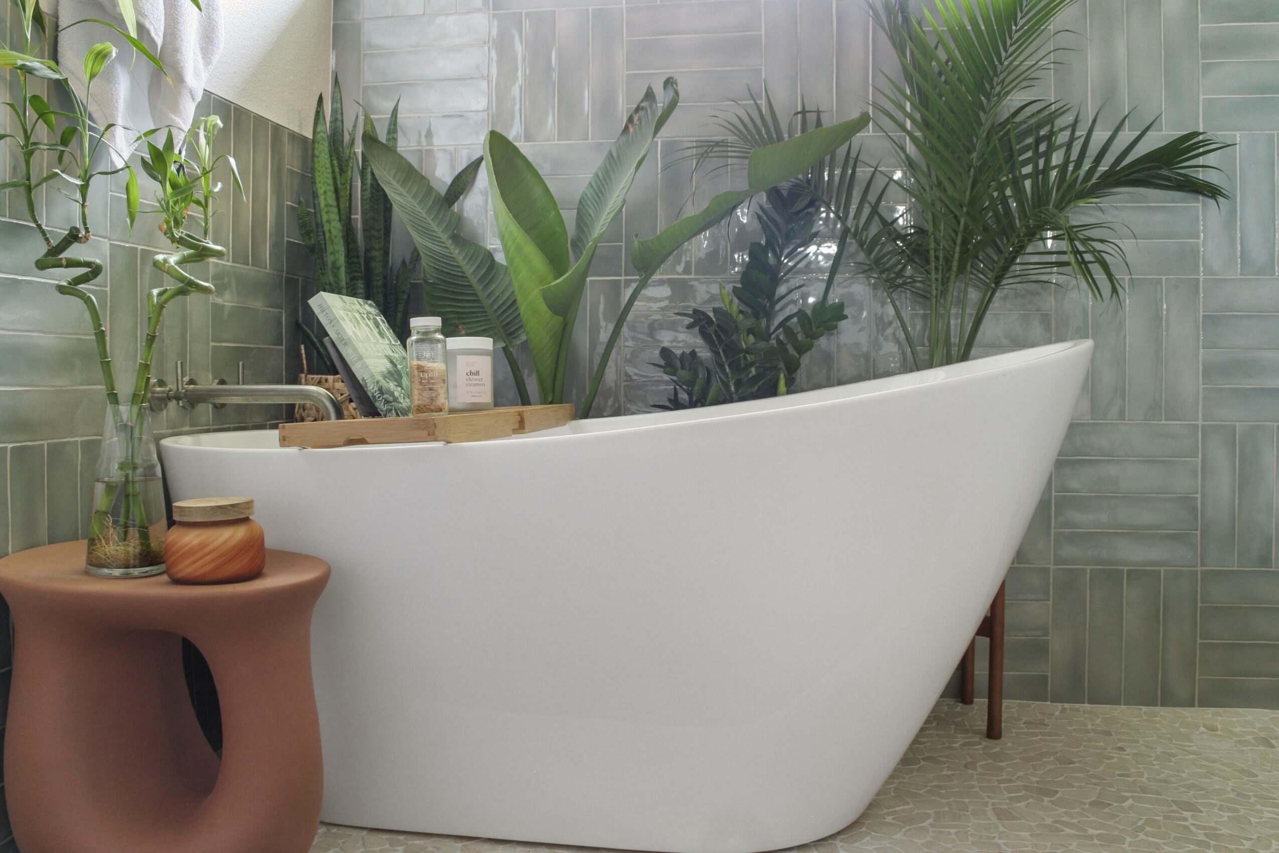 White bathtub in a bathroom with green plants and textured gray wall tiles, next to a small pink side table.