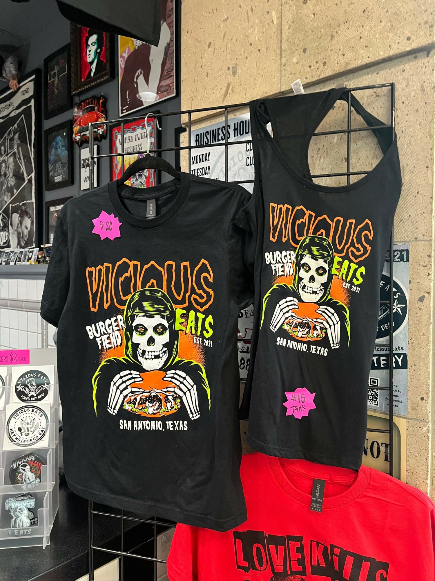 Two black sleeveless shirts with skeleton and skull designs, hanging on a metal rack, with a red bag nearby.