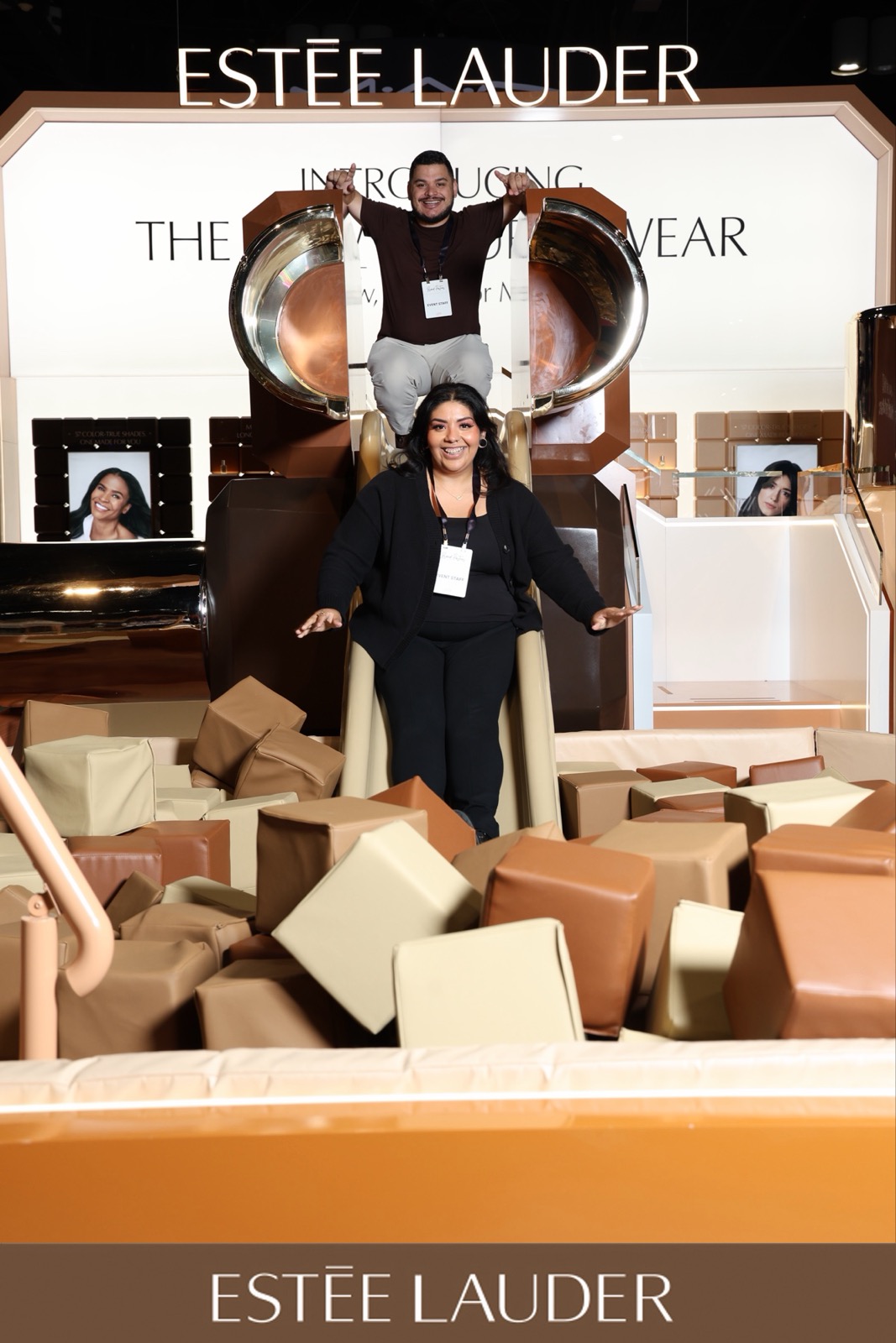 Two women stand in front of a large Estée Lauder display with makeup products and boxes, one woman is on a platform, surrounded by boxes.