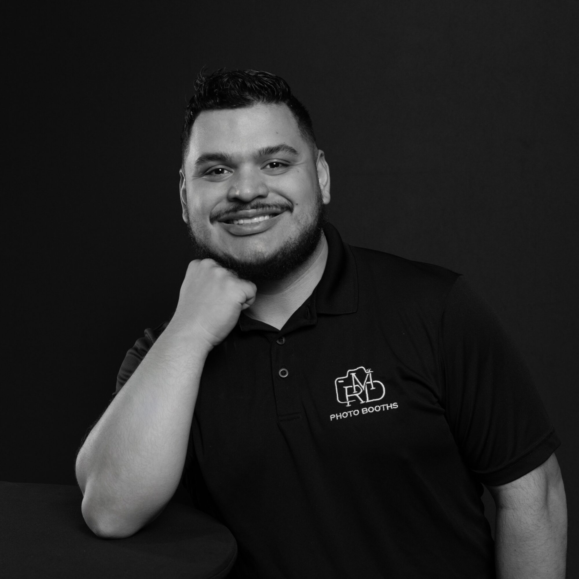 Man with short hair and beard smiling, resting chin on hand, wearing a polo shirt with logo, against dark background.
