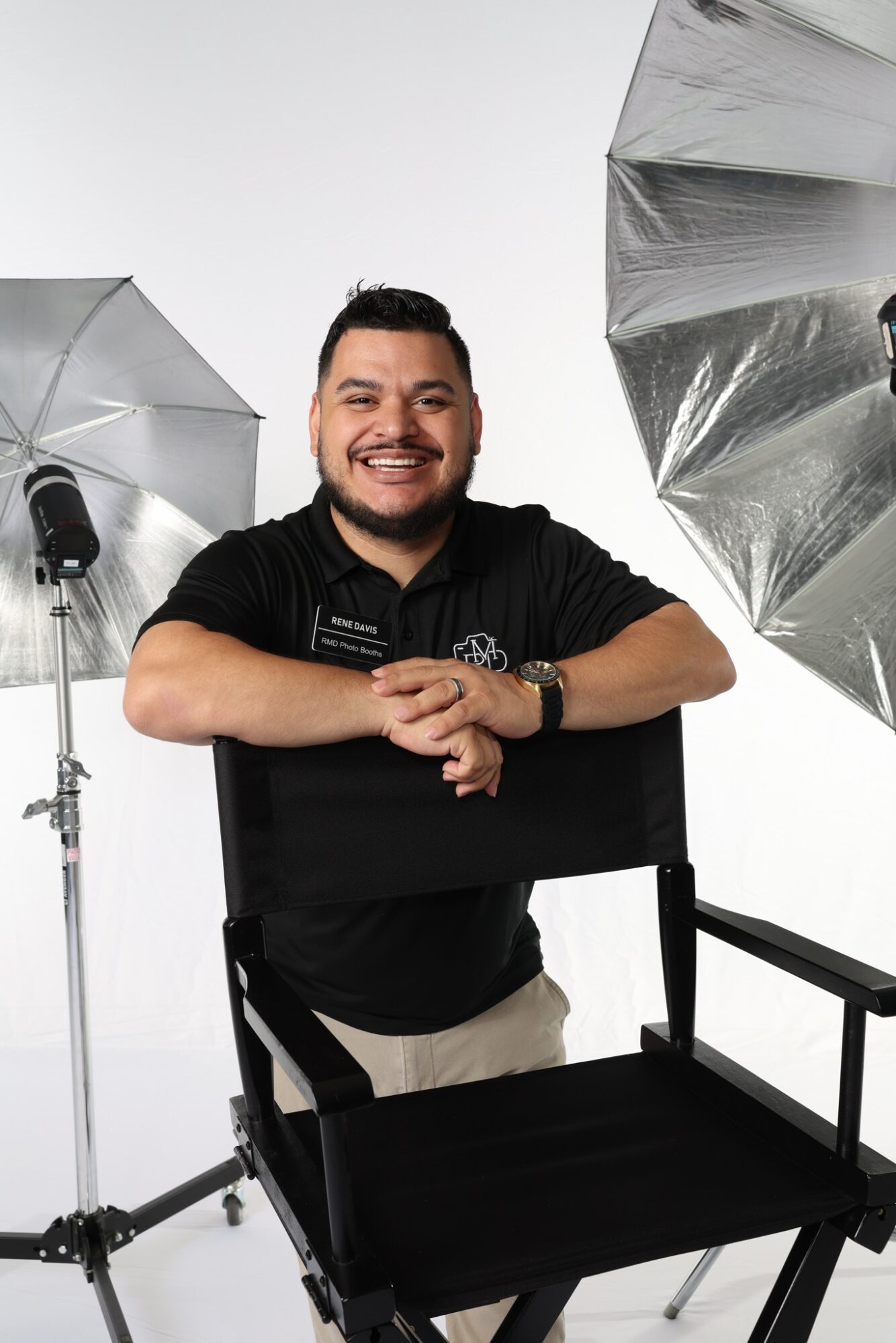 Man smiling, sitting behind a black chair, with photography umbrellas and lights in the background.
