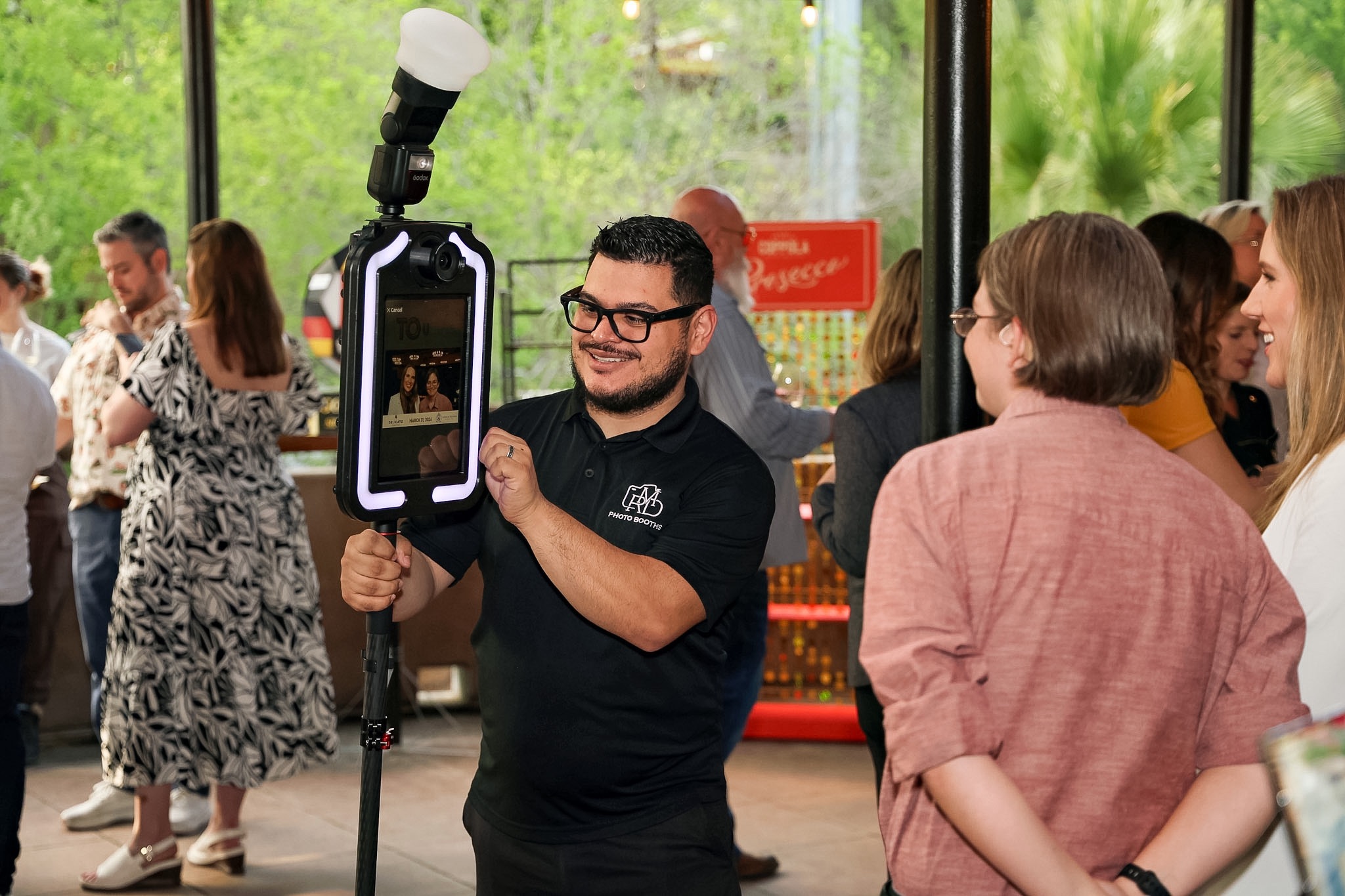 Man with glasses and a black shirt takes a photo with a smartphone and ring light, surrounded by people indoors.