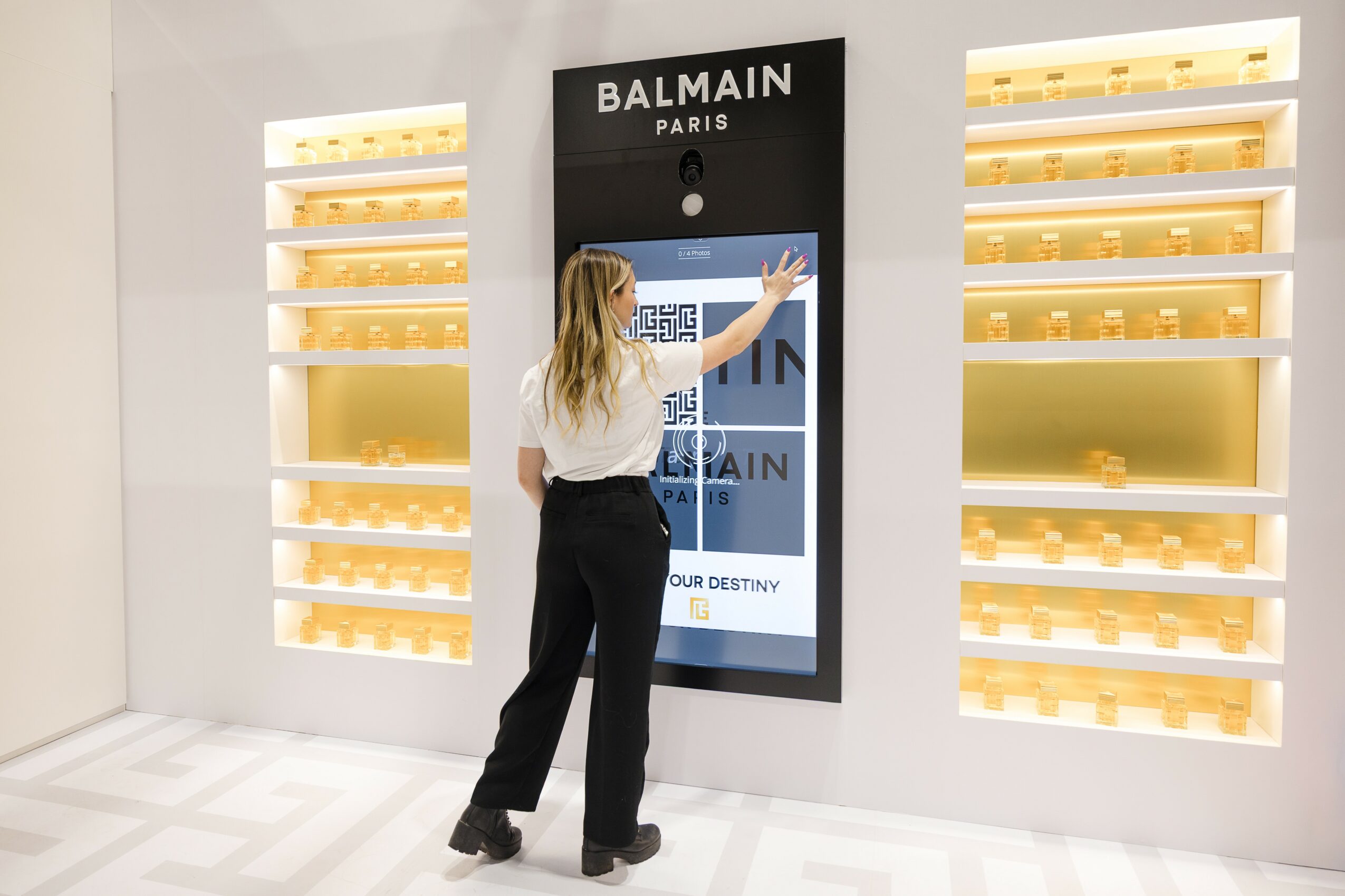 Woman interacts with a digital display at a Balmain Paris store, with shelves on either side, in a modern setting.