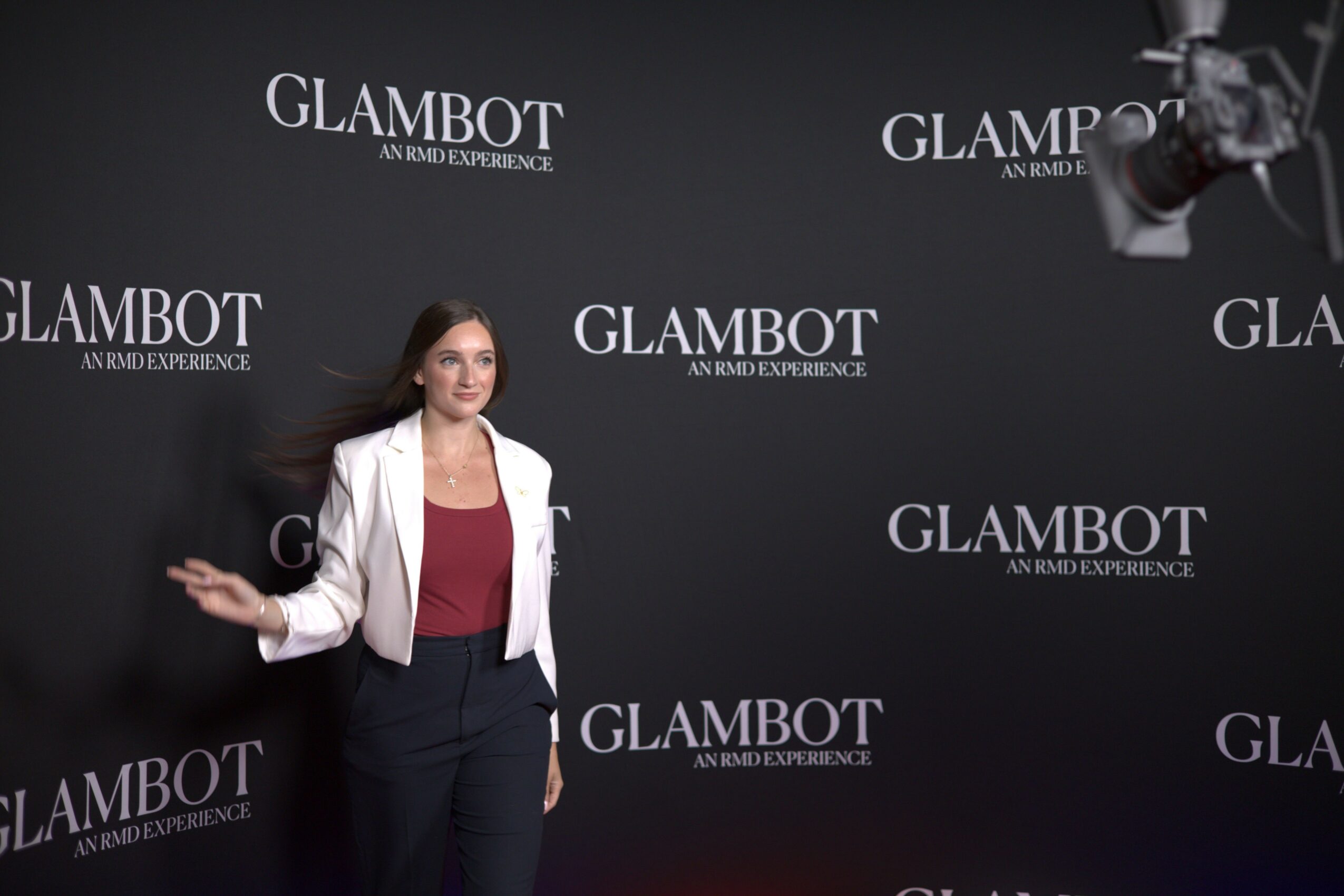 Woman standing in front of a black backdrop with 'GLAMBOT' logos, wearing a white blazer and dark pants.