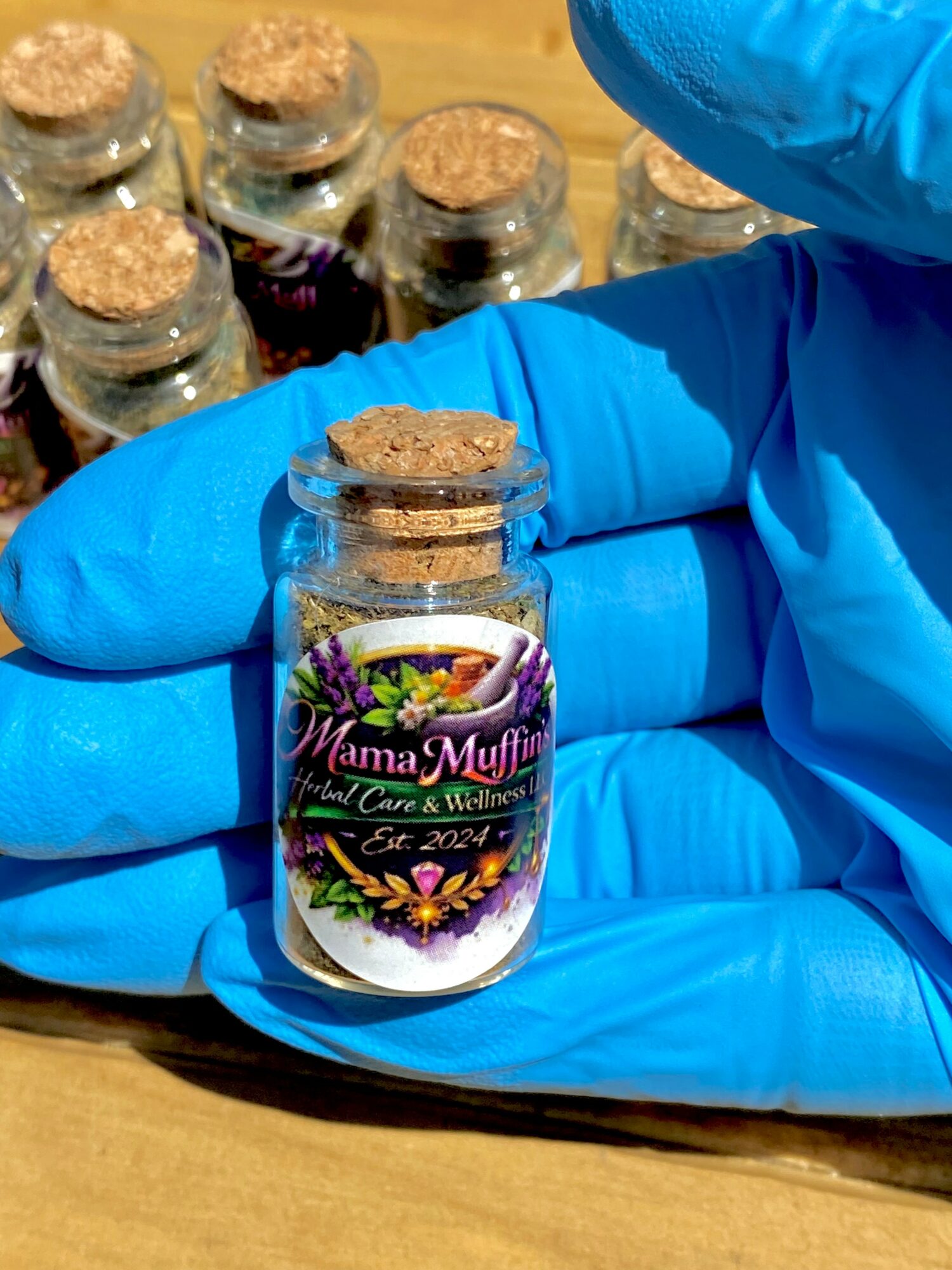 Person wearing blue gloves holding a small glass jar with a colorful label, cork lid, and other jars in background.