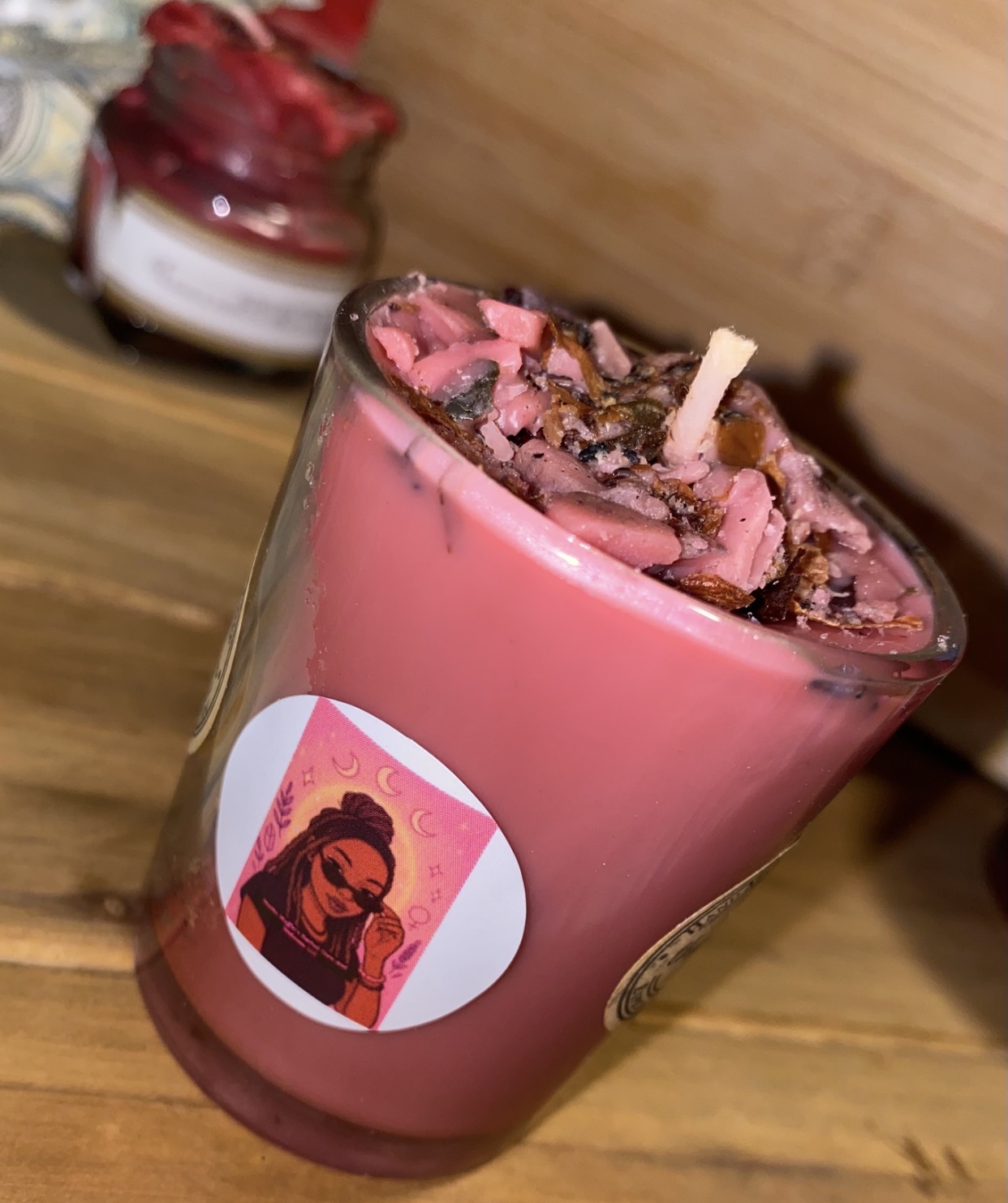 Pink candle with a label of a woman with a headscarf, topped with pink and brown decorative pieces, on a wooden surface.