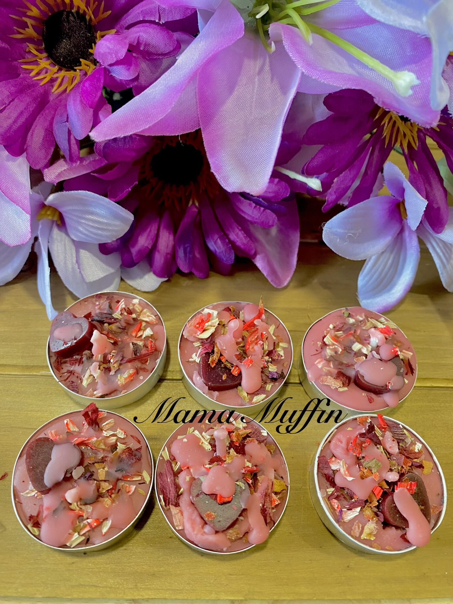 Six small candles with pink and red flower petals on a wooden surface, with a bouquet of purple and pink flowers in the background.