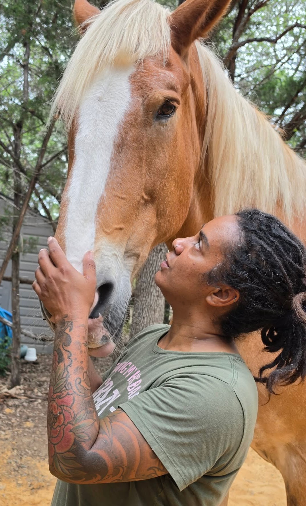 Woman with tattoos gently touches a horse's face outdoors, trees in background.