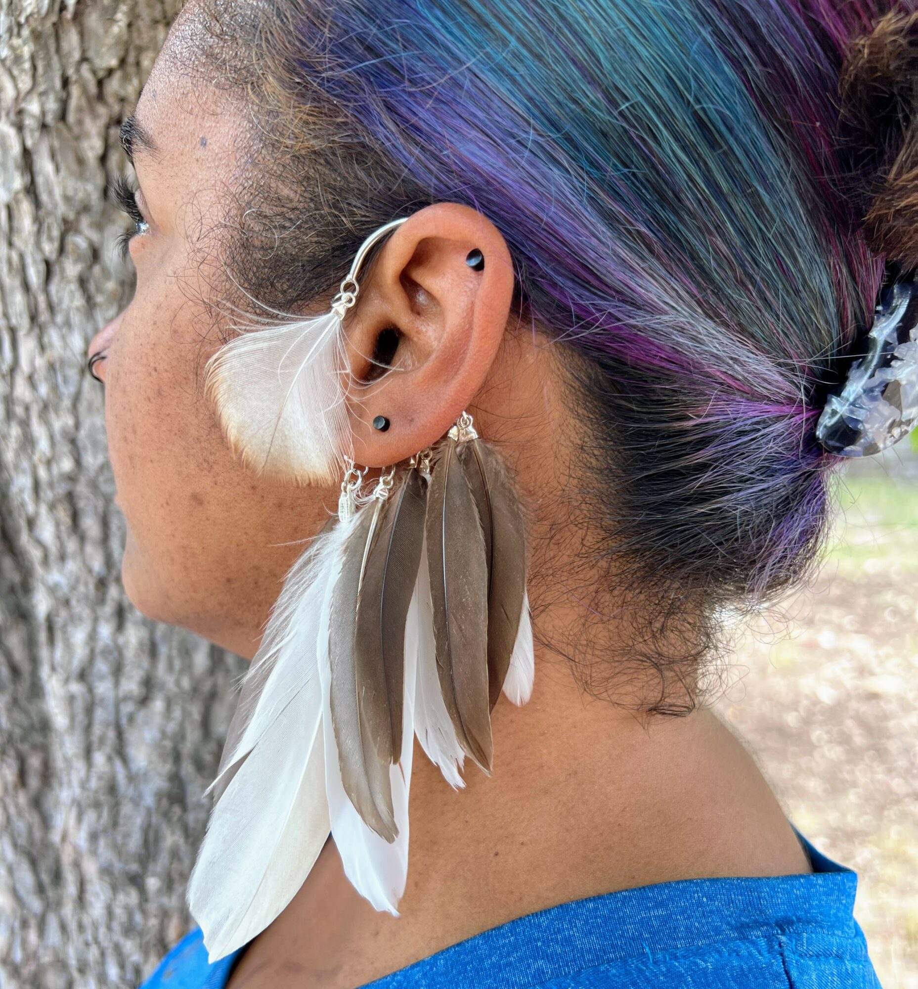 Side view of a person with colorful hair, multiple earrings, and large feather earrings, standing outdoors near a tree.