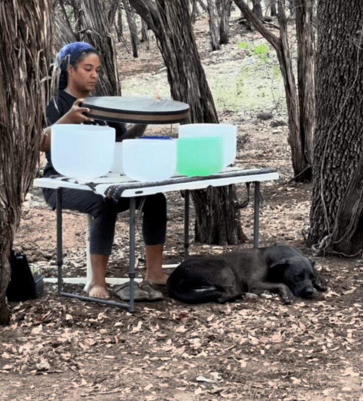 Girl with long hair and headband playing a drum set outdoors, with a black dog lying on the ground nearby, surrounded by trees.